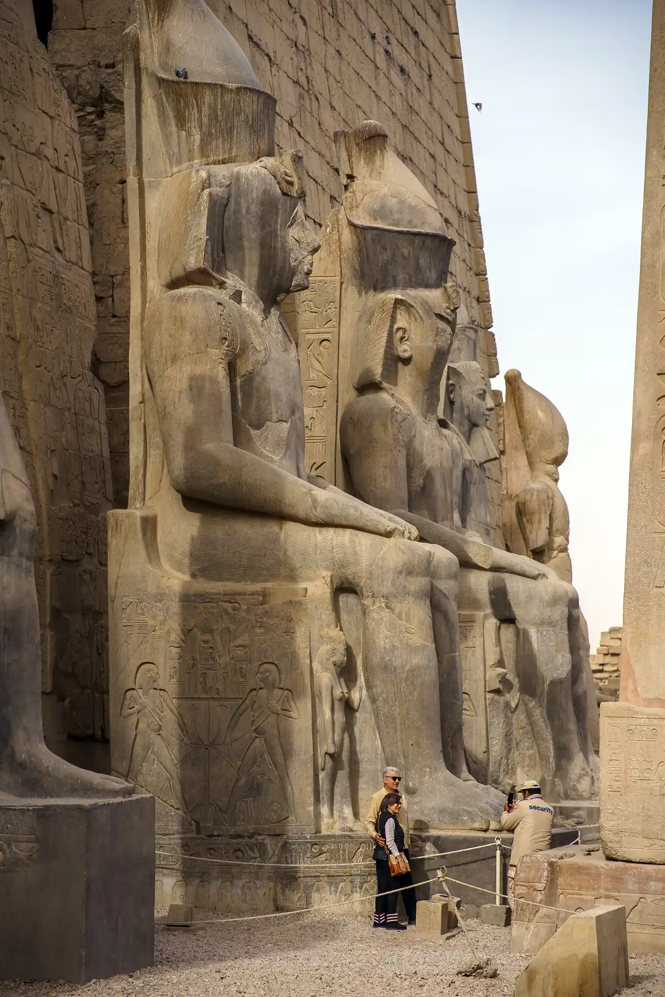 Colossal seated pharaoh statues tower over two visitors at the entrance of Luxor Temple, their carved bases bright in the sun.