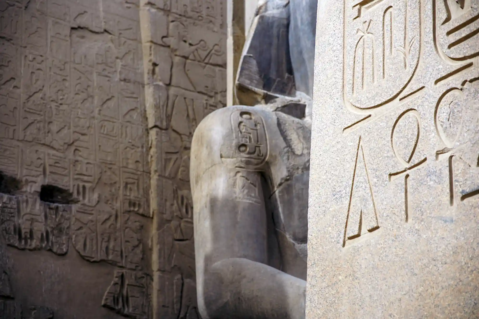 A pharaoh statue stands beside sunlit walls etched with hieroglyph reliefs at Luxor Temple.