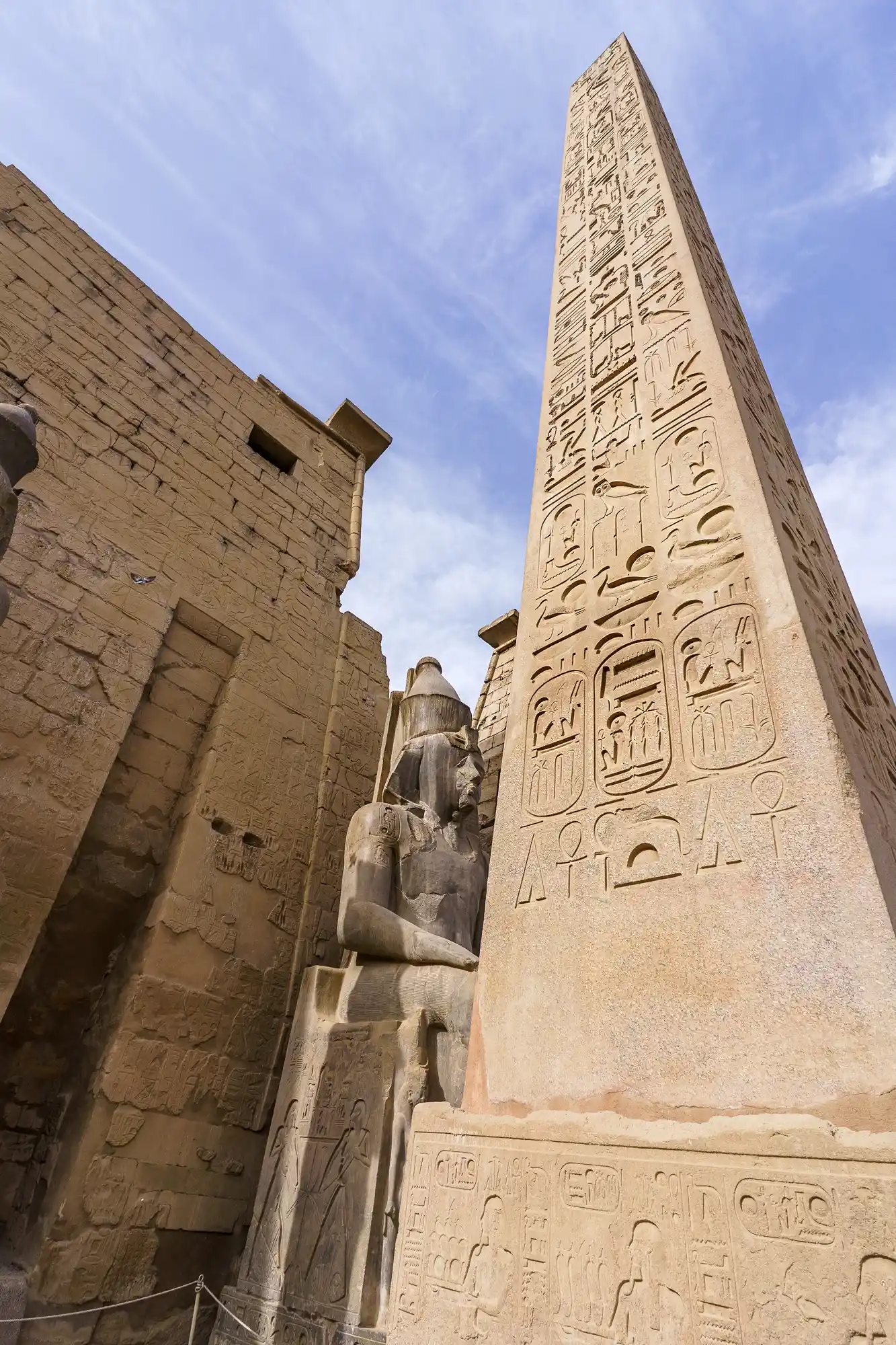 A seated pharaoh statue beside the hieroglyph-covered obelisk at Luxor Temple, set against a pale blue sky.