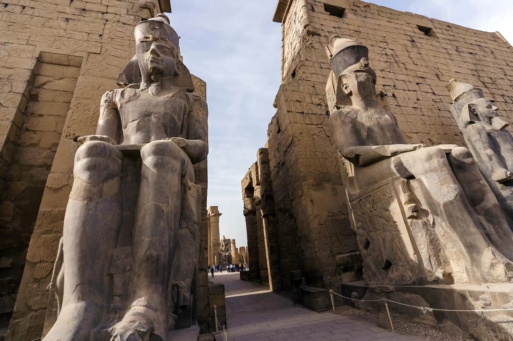 Seated stone pharaohs line the way into Luxor Temple in low sunlight, with tiny visitors far in the distance.