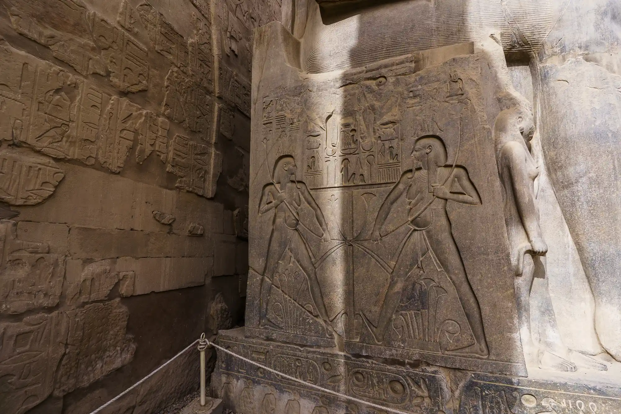 Two figures carved in a sandstone relief at Luxor Temple, lit from above, with a rope barrier keeping visitors back.