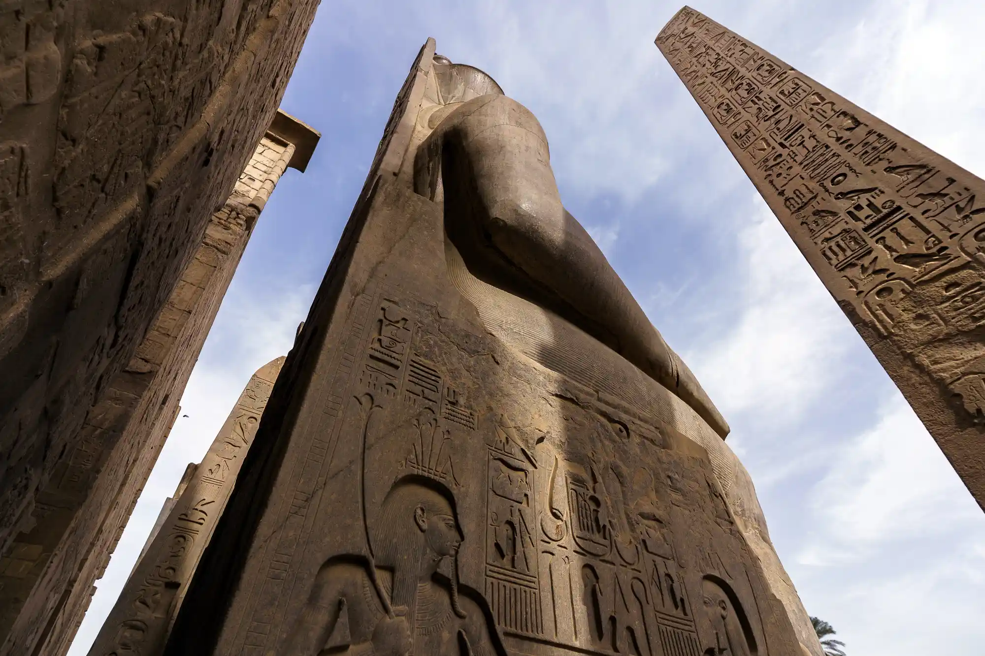 A carved pharaoh statue rises against a pale blue sky at the entrance to Luxor Temple, figures etched into its base.