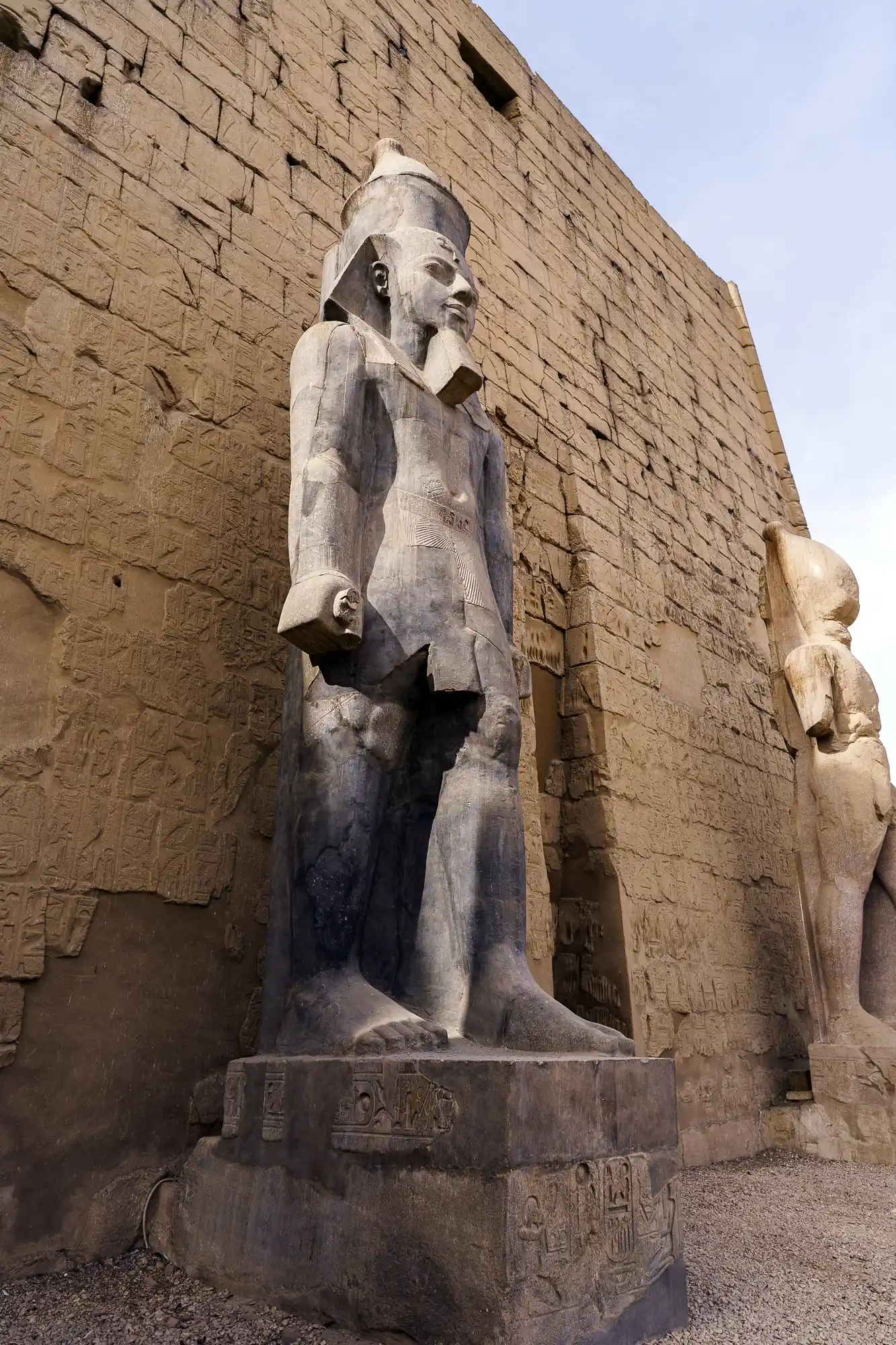 A sandstone pharaoh statue in Luxor stands in bright sun, casting a sharp shadow on the tan blocks behind it.