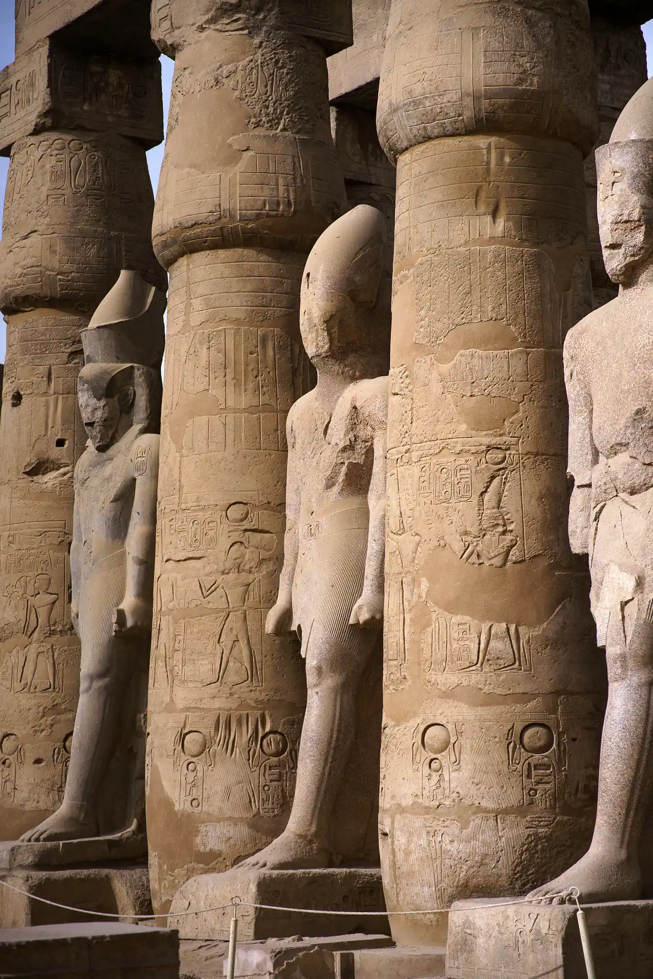 Standing pharaoh statues in front of carved columns at Luxor Temple, lit from directly above.