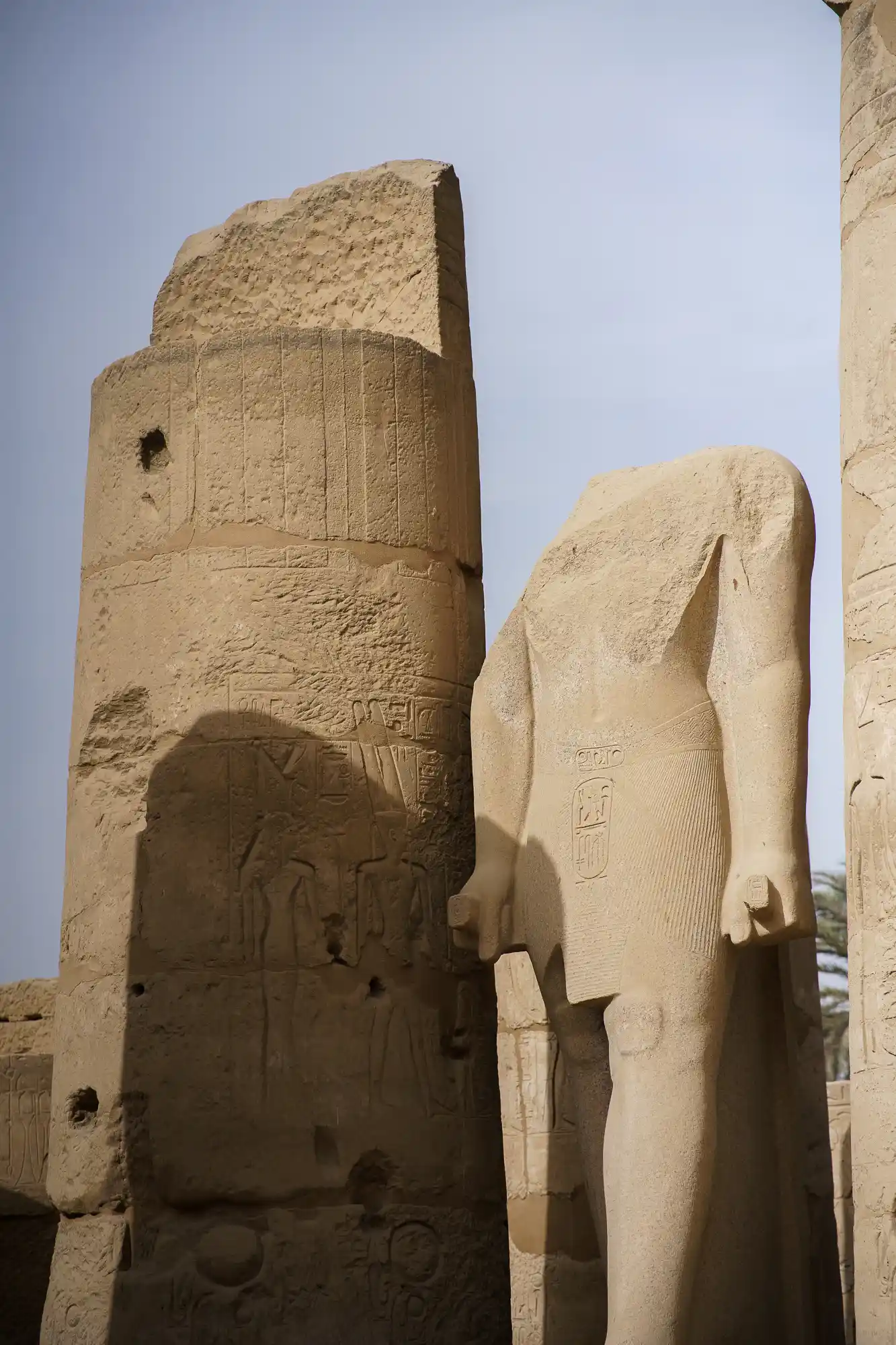 A headless beige sandstone statue beside a carved pillar at Luxor Temple, side-lit so every chisel mark shows.