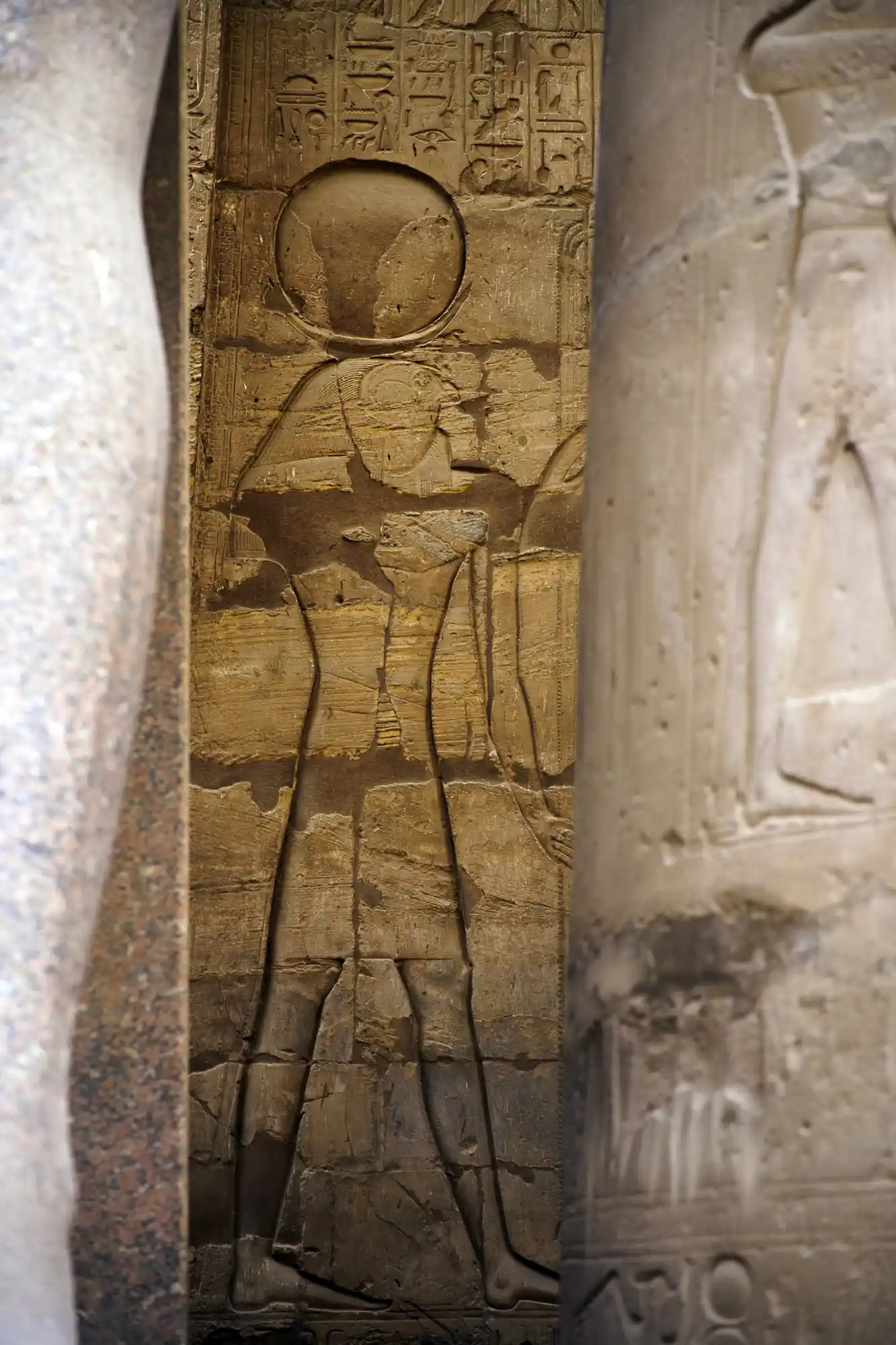 A worn sandstone relief in Luxor Temple shows a figure crowned with a sun disk, framed by pale stone pillars.