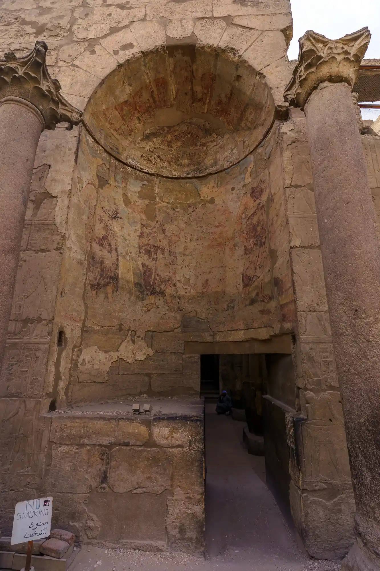 Carved columns frame a shadowy niche with traces of faded red paint at Luxor Temple, a visitor sitting inside.
