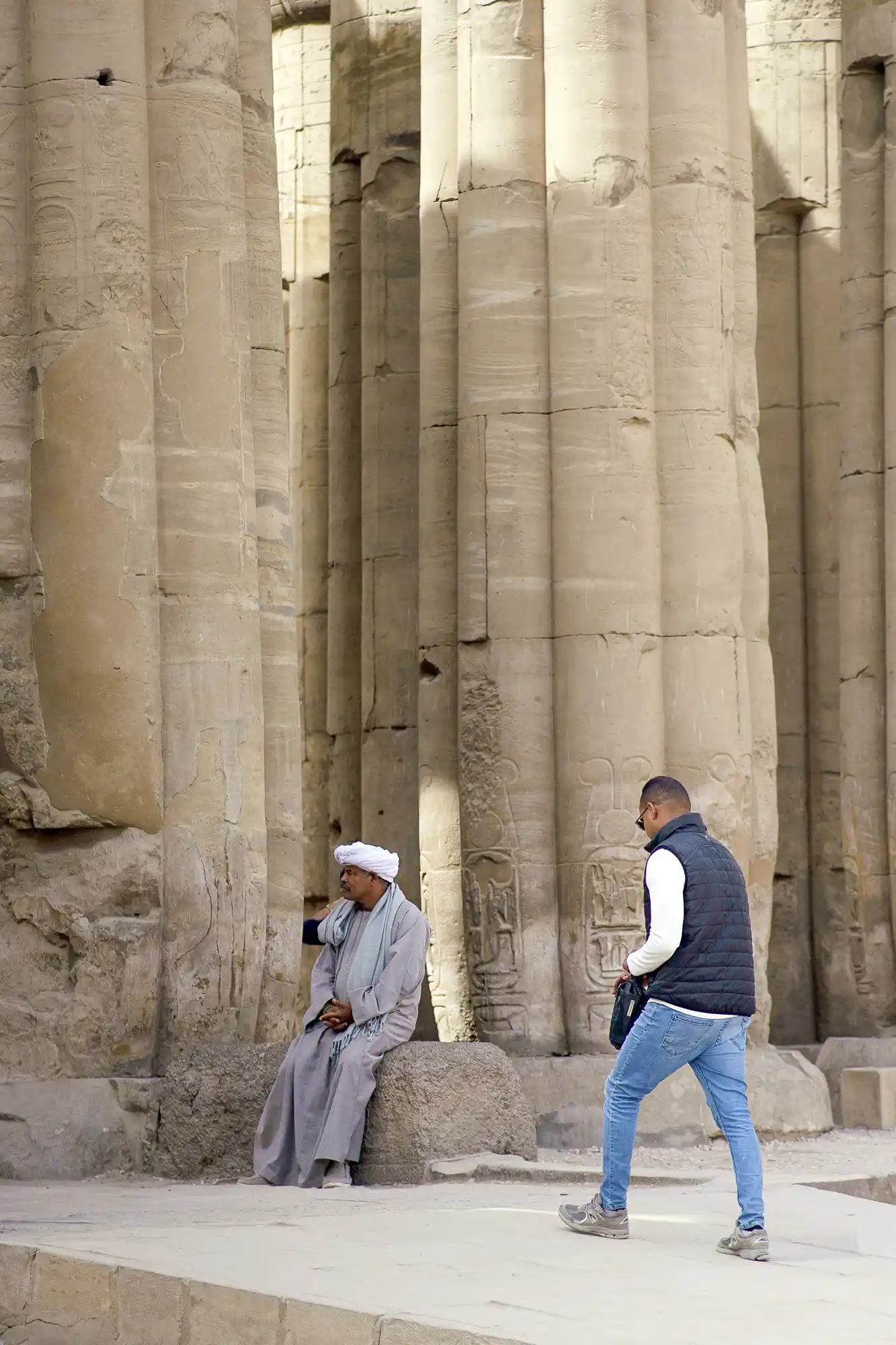 A man in a grey robe rests on a stone block as another walks past the carved papyrus columns of Luxor Temple.