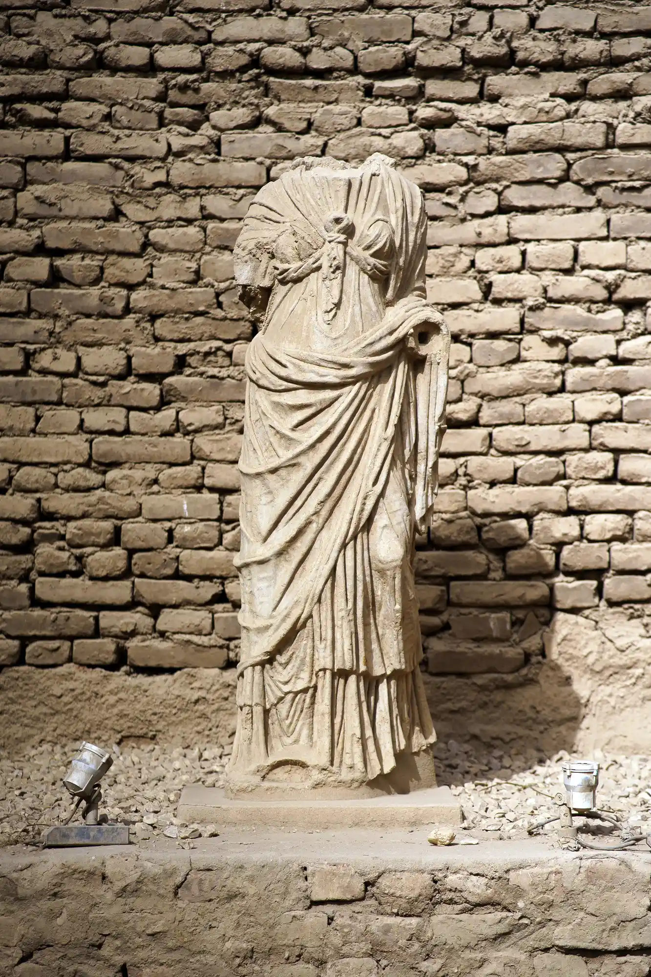 A headless statue in flowing draped robes stands before a rough brick wall at Luxor Temple, lit by two angled lamps.