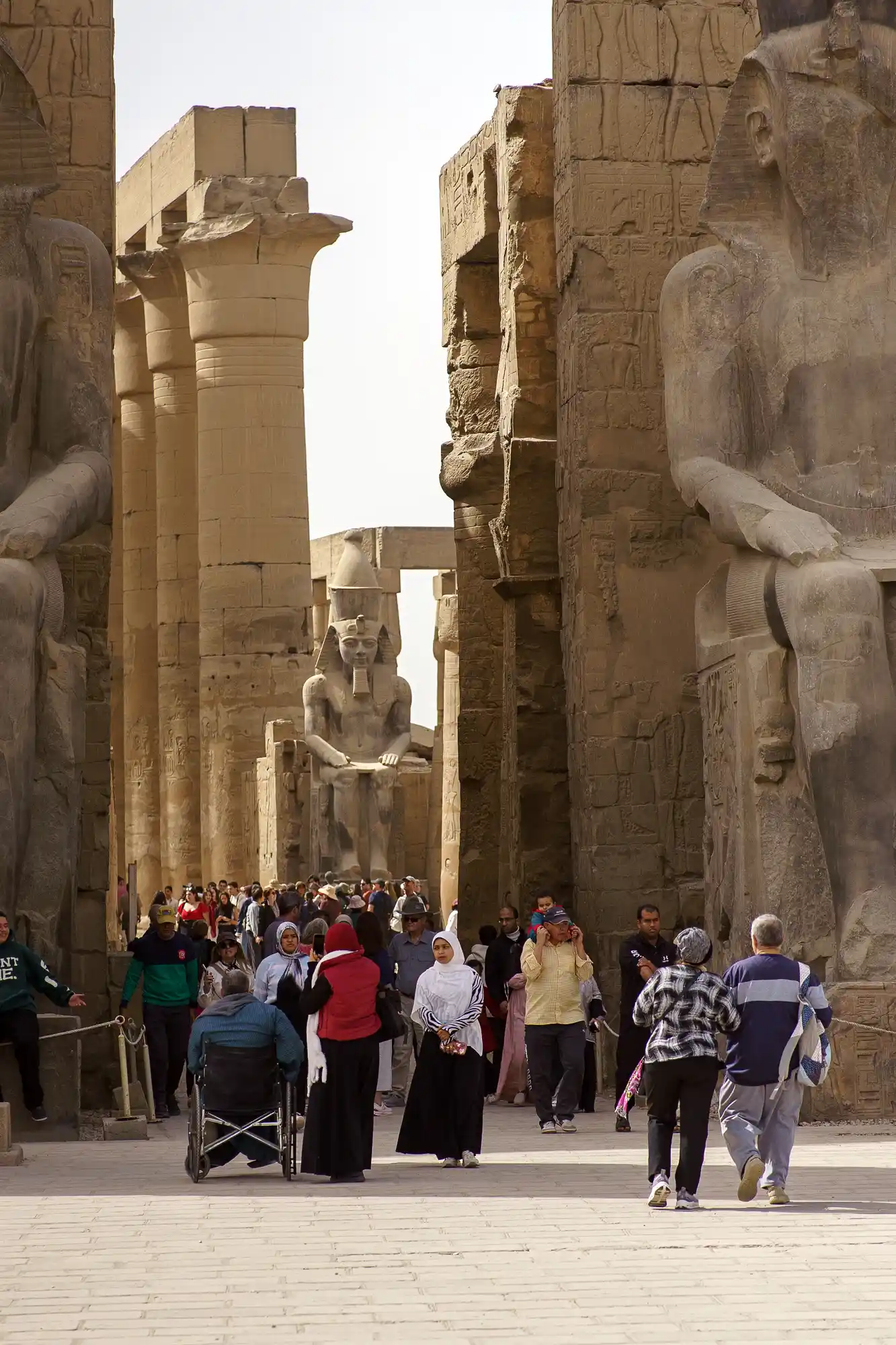 A crowd of visitors, including a wheelchair user, moves between seated statues and sunlit columns at Luxor Temple.