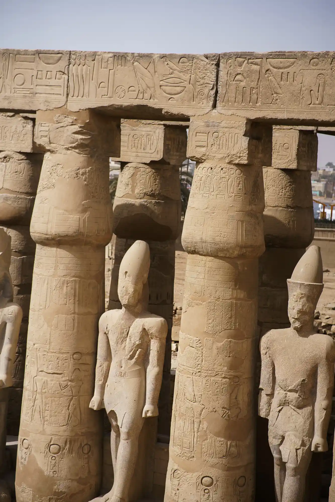Stone pharaoh figures stand among hieroglyph-carved columns at Luxor Temple in bright overhead light.
