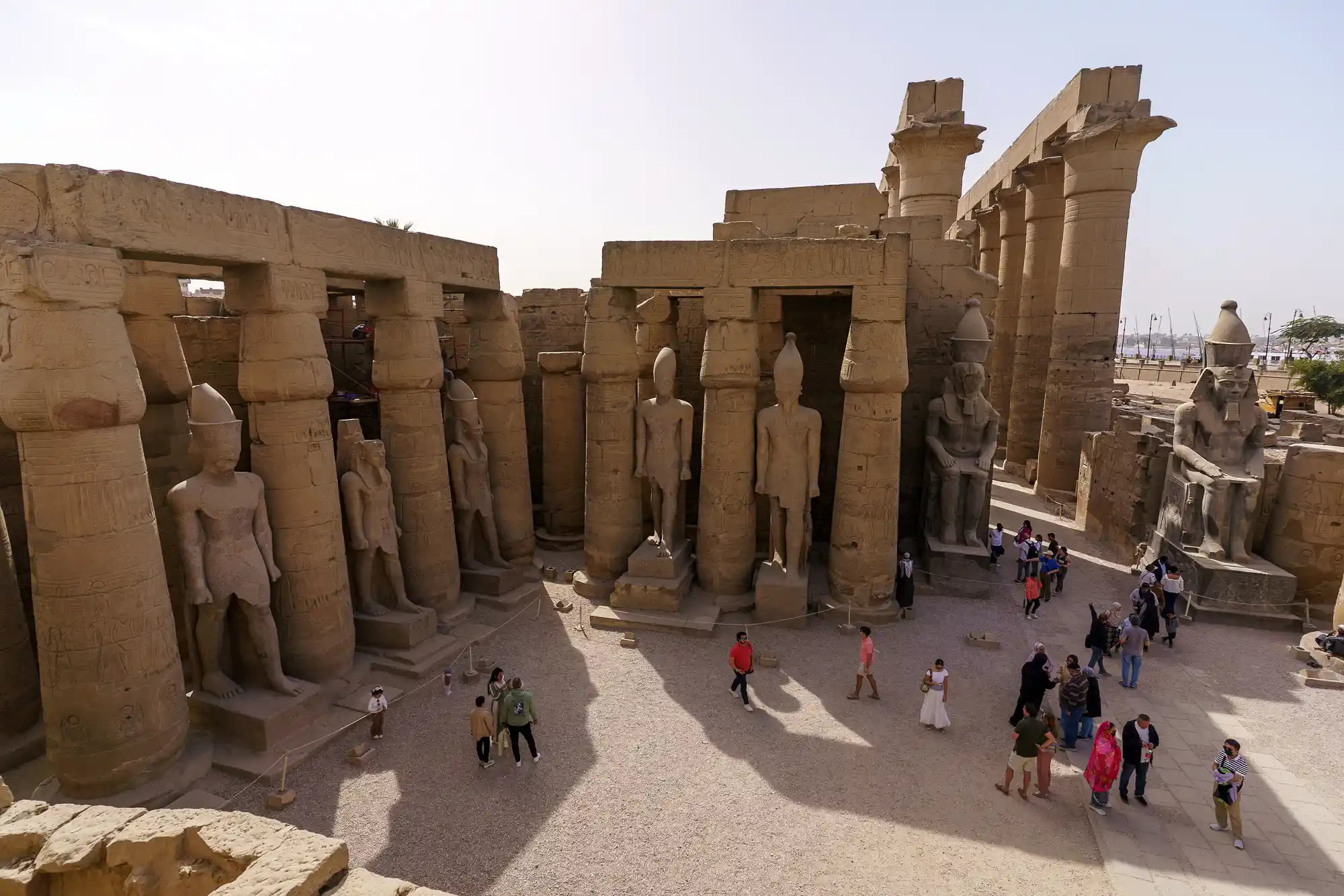 Pharaoh statues cast long shadows across visitors on a sunny day at Luxor Temple.