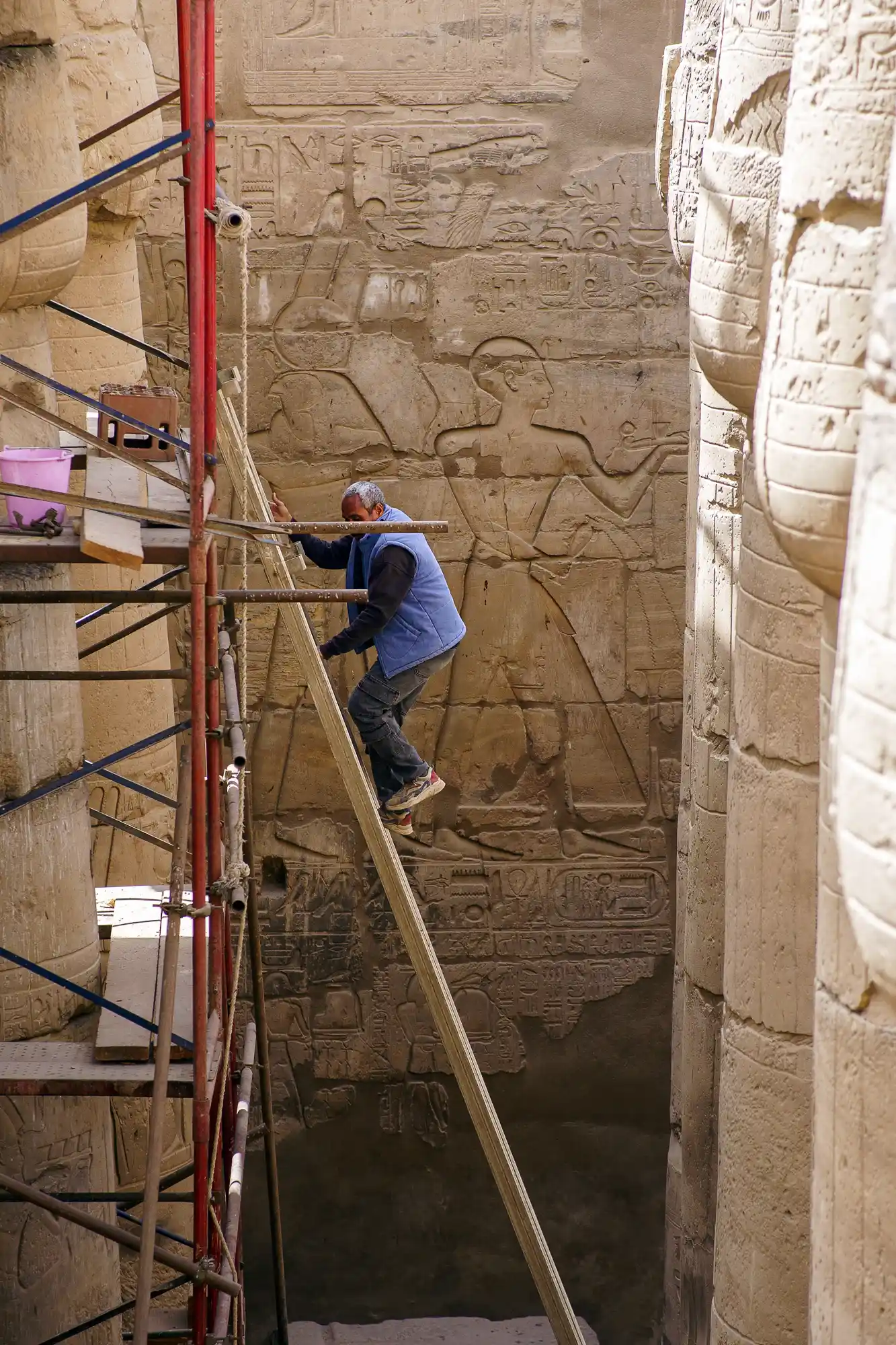 A worker in a blue vest climbs a wooden plank beside red metal scaffolding during restoration work at Luxor Temple.