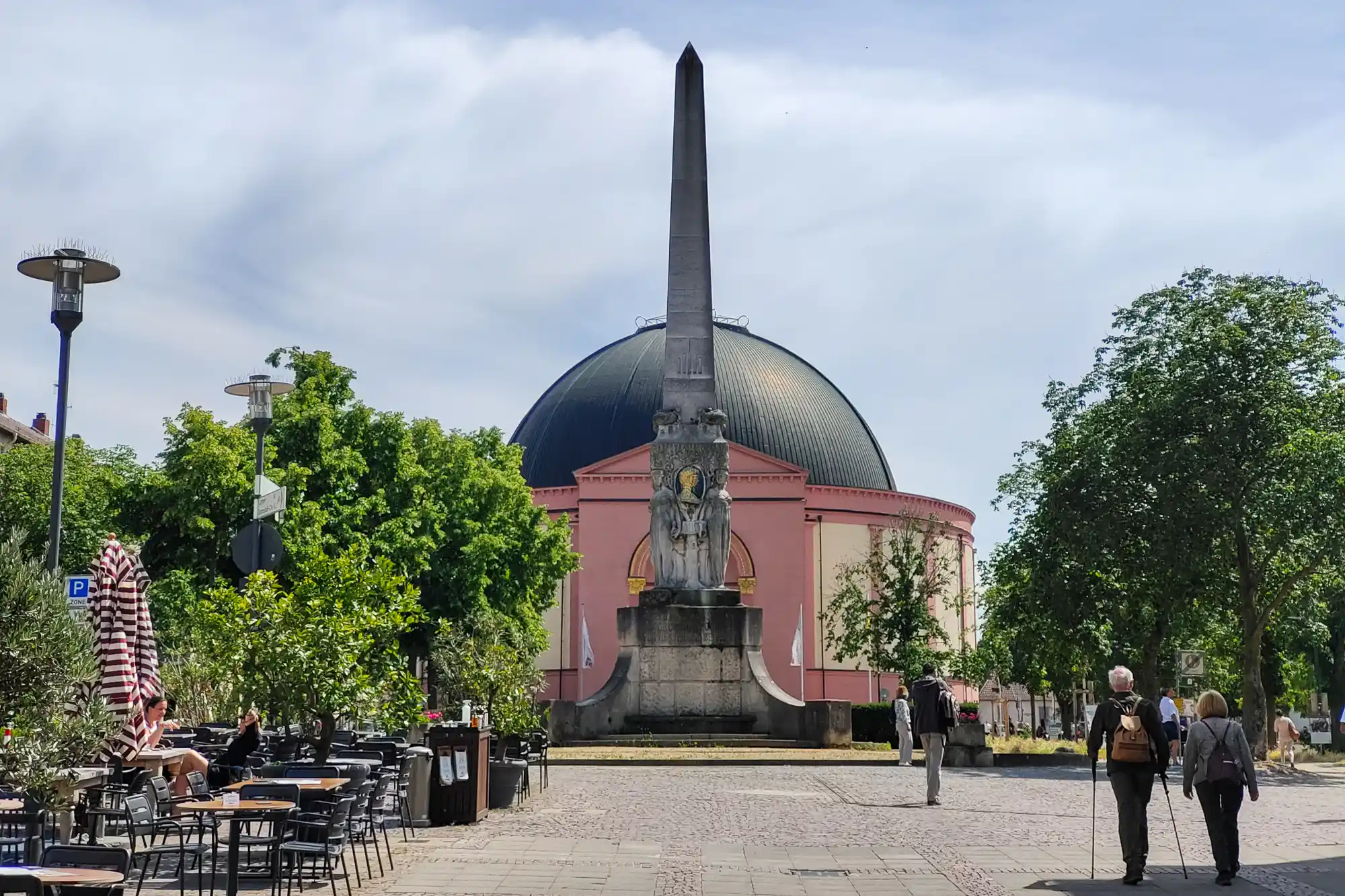 A stone obelisk with carved figures in front of the pink St. Ludwig's Church and its dark dome in Darmstadt, with people crossing the square.