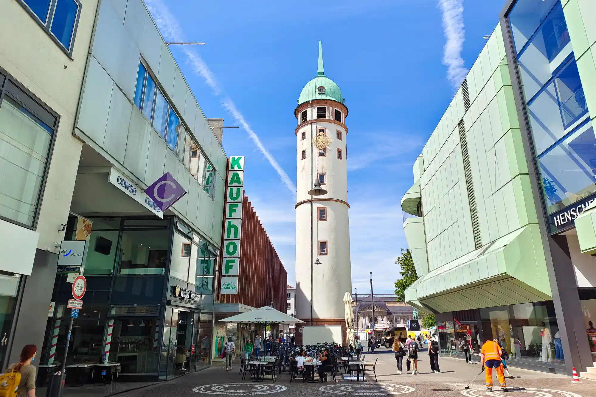 The Weisser Turm, a white tower with a green dome, rises above a paved square in Darmstadt, with people walking past glass buildings.