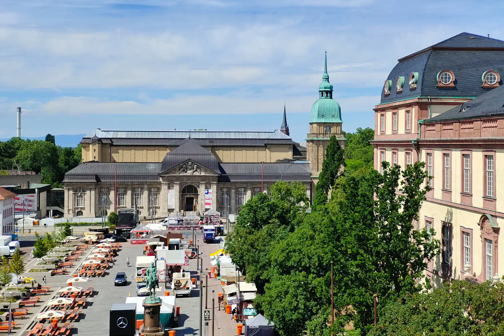 A tan stone building with a green dome in Darmstadt in bright light, with orange tables and green trees in the foreground.
