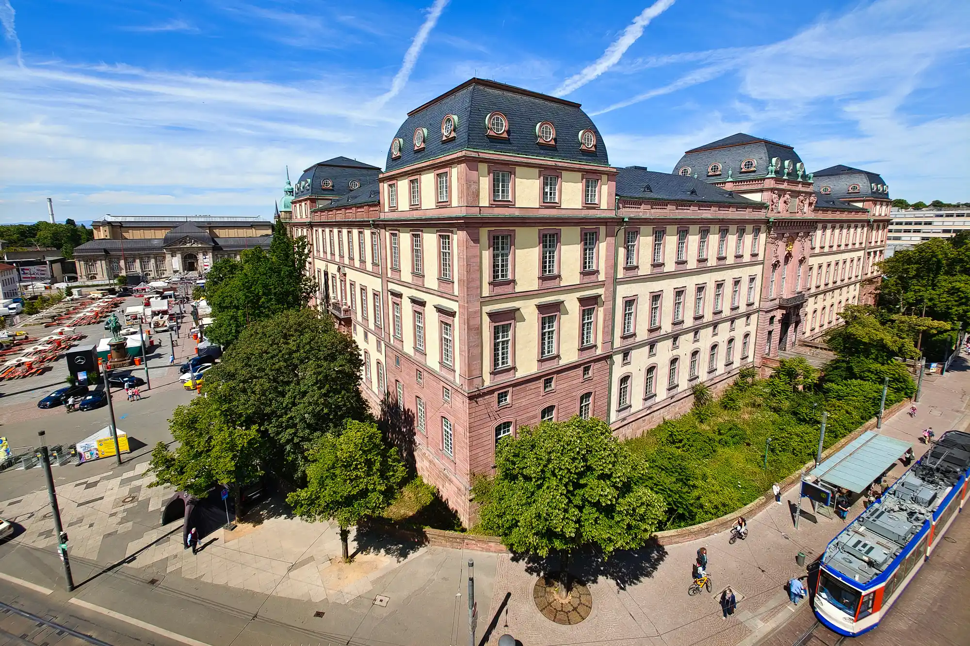 A tan and red building with a dark roof in Darmstadt in bright light, with green trees and a blue and white tram below.