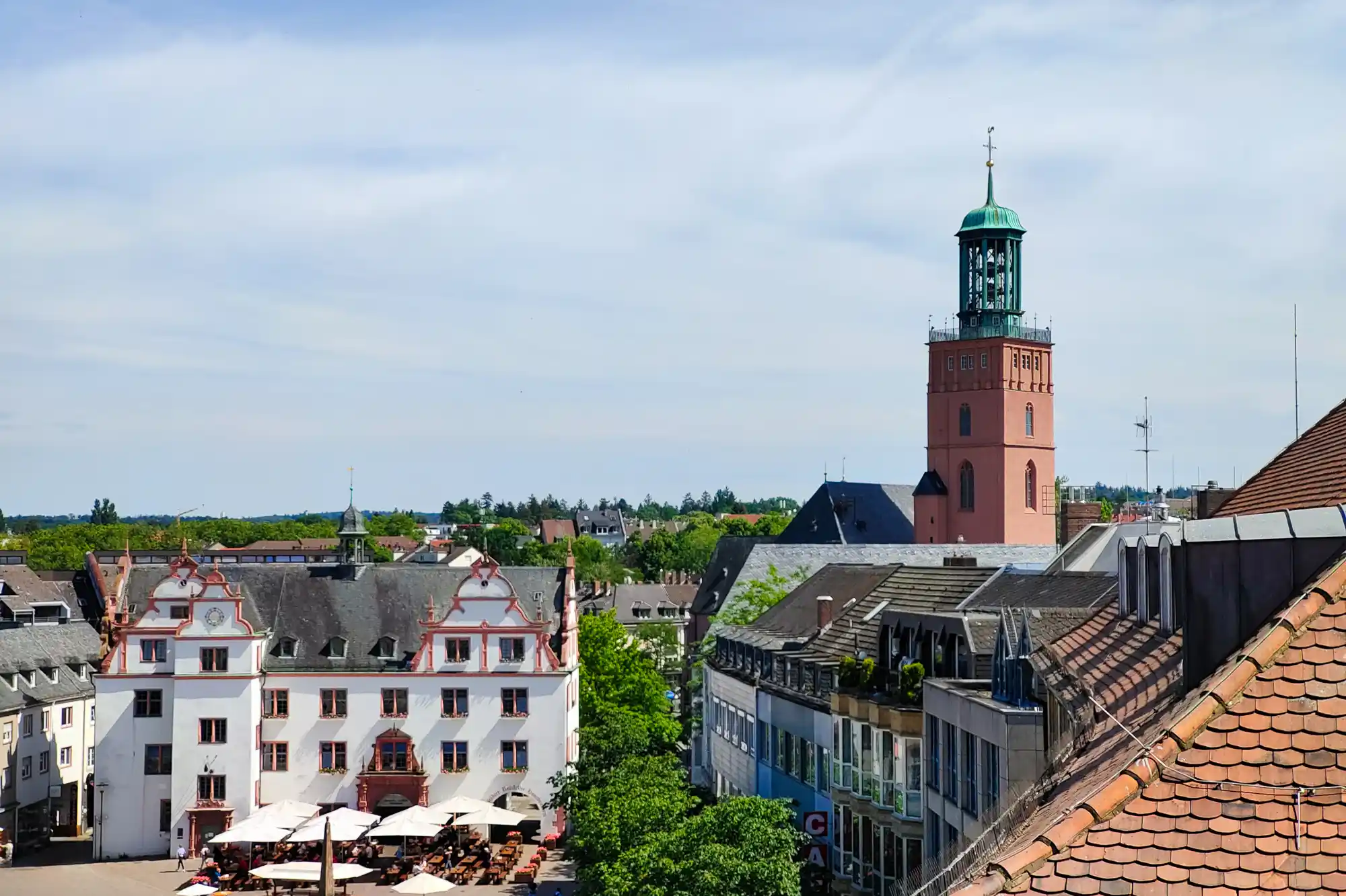 A pink tower with a green top rises above buildings in Darmstadt, while white umbrellas shade people below in bright light.