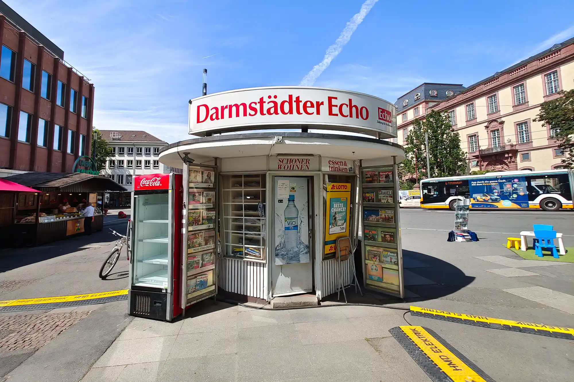 A white kiosk with red lettering and a red cooler on a Darmstadt street, with yellow strips marking the ground in bright light.