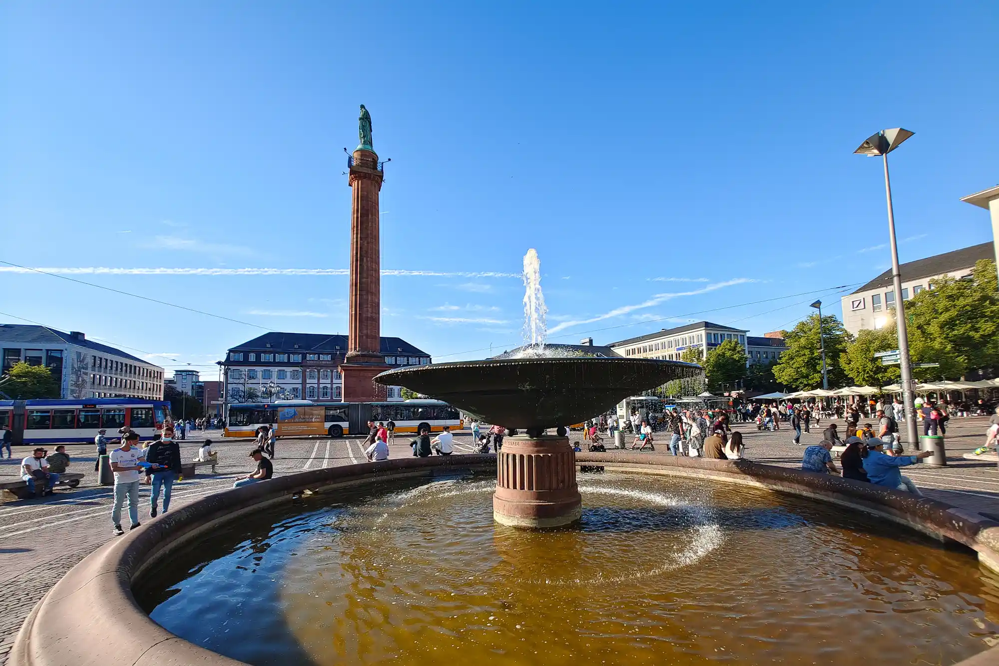 A dark fountain sprays water in bright sunlight in Darmstadt, with a red column and people standing nearby.