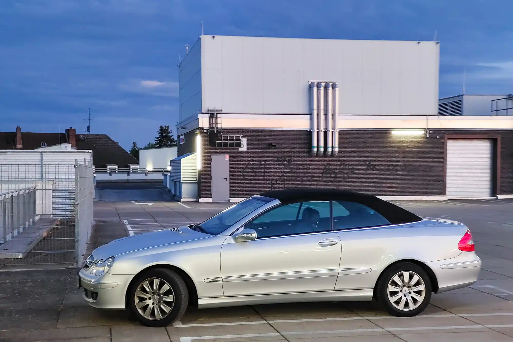 A silver convertible with a black top parked on concrete in Darmstadt, blue evening light reflecting off its windows by a brick wall.