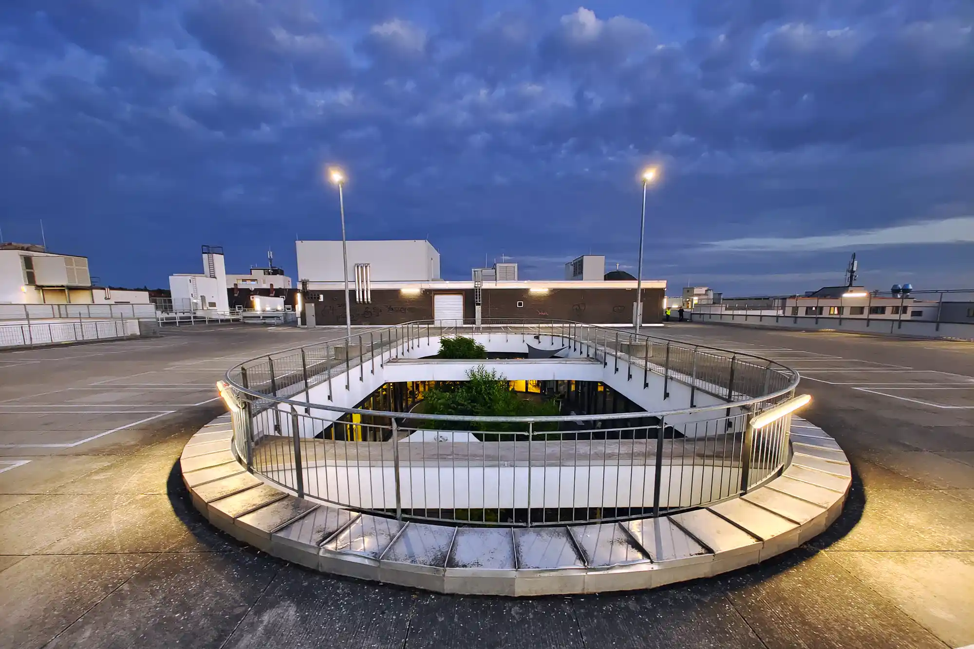 A circular metal railing around a sunken opening with green plants in Darmstadt, the concrete lit by warm lights under a blue sky.