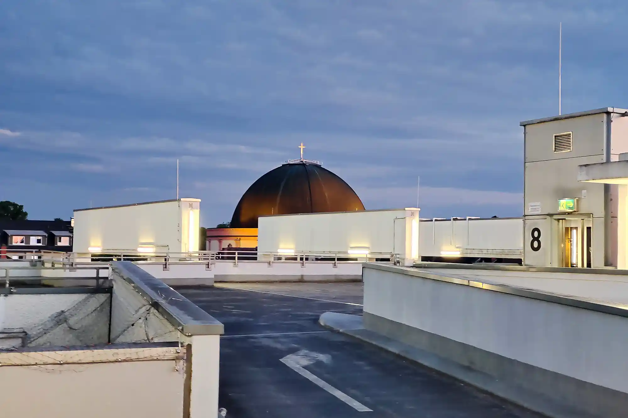 The dark dome and cross of St. Ludwig's Church, the Käseglocke, rise behind white concrete walls in Darmstadt under a blue evening sky.