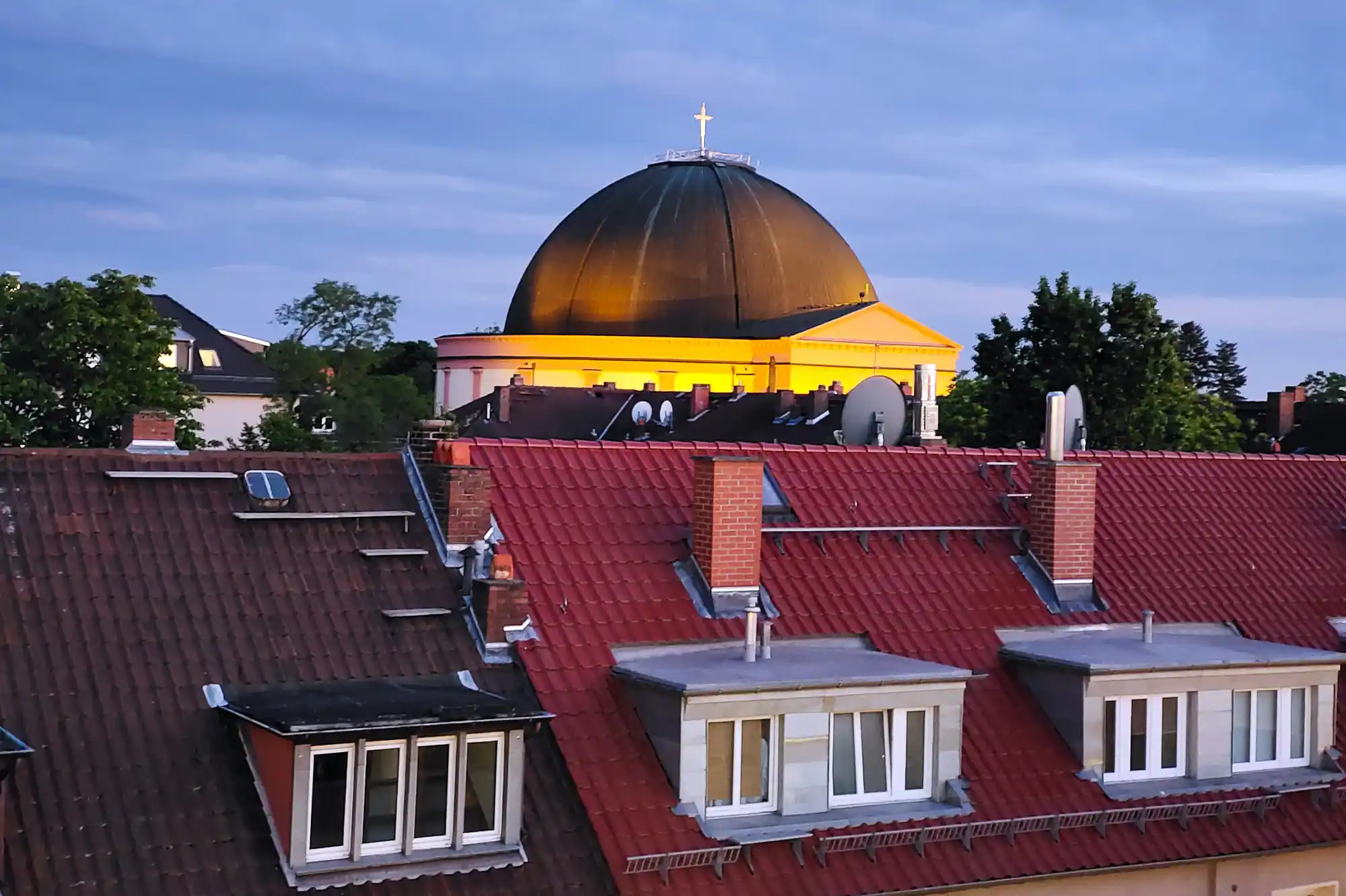 The dome of the Ludwigskirche glowing gold above red tile roofs in Darmstadt, framed by brick chimneys and green trees in the evening.