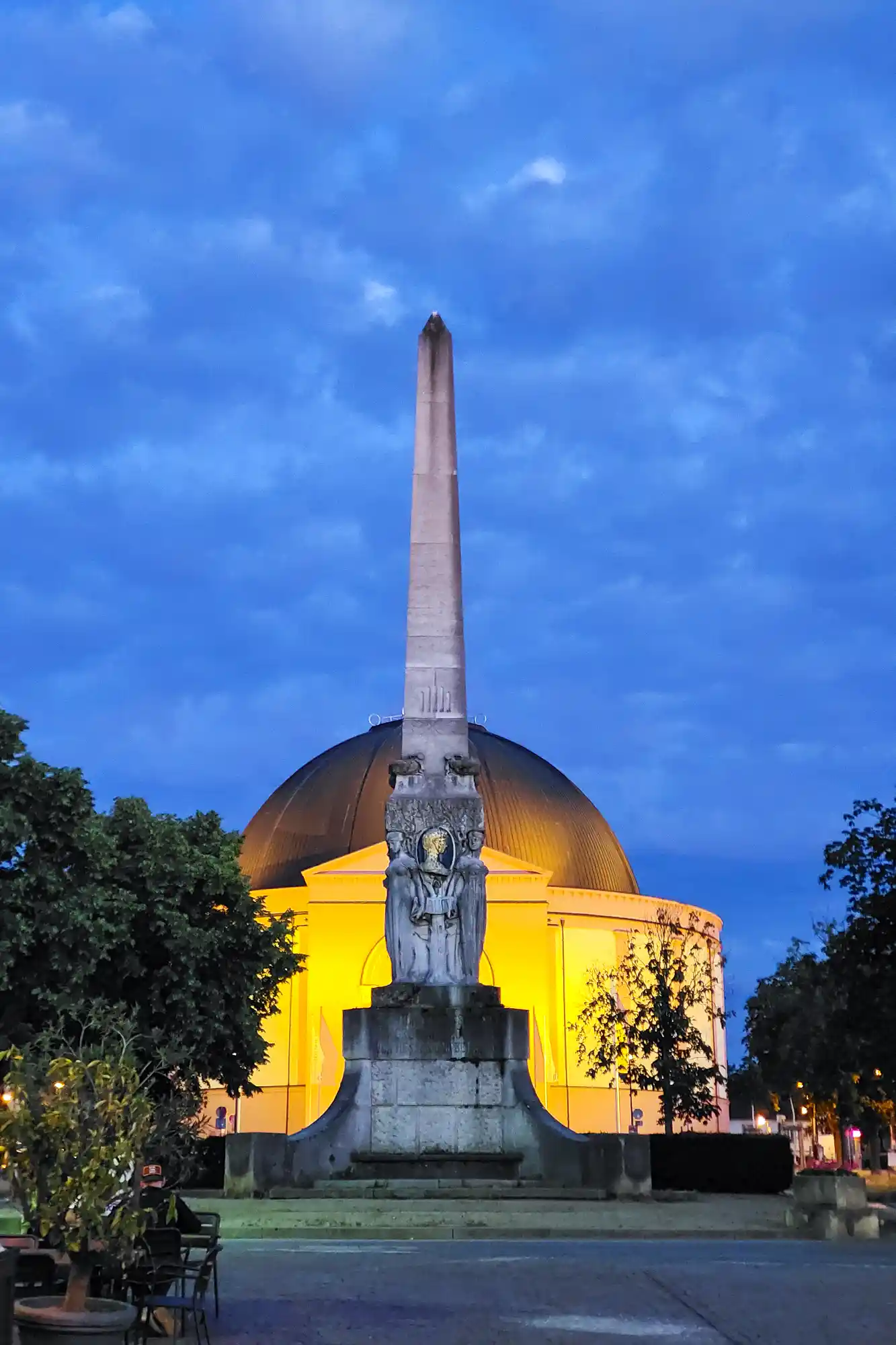 A carved stone obelisk at dusk in Darmstadt, standing in front of the yellow-lit dome of St. Ludwig's Church under a deep blue sky.