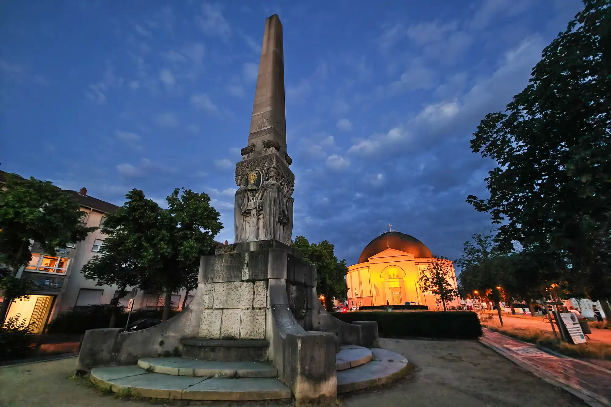Low-angle view of the carved stone obelisk in Darmstadt, with a domed building glowing in warm yellow light behind it.