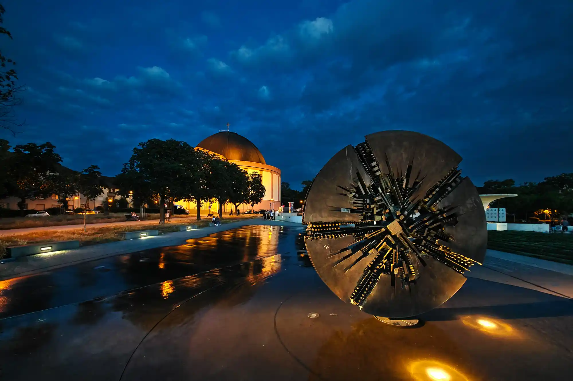 A dark spiky metal sculpture on wet ground reflecting warm light in Darmstadt, with the Ludwigskirche lit up under a deep blue sky.
