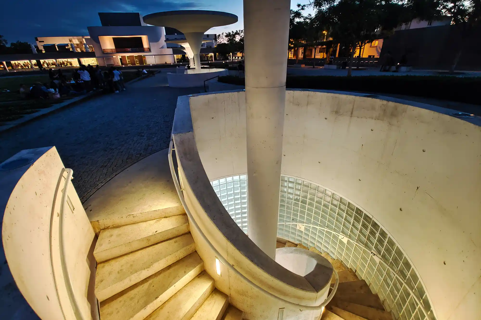 A spiral concrete staircase winds down around a pillar in Darmstadt, lit by warm light, with glass blocks lining the curved wall.
