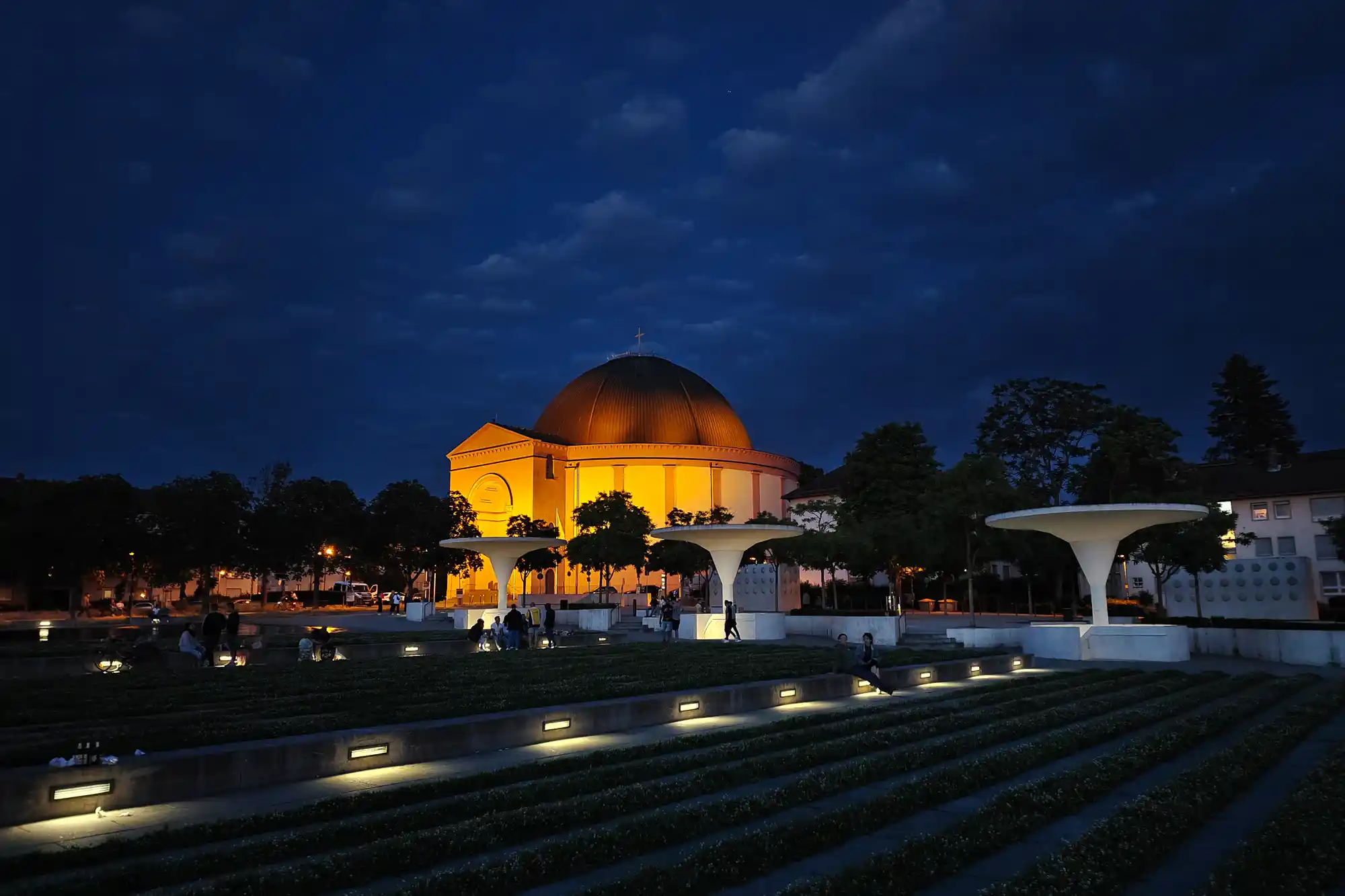 The glowing yellow dome of St. Ludwig's Church in Darmstadt under a dark blue sky, with people sitting on tiered grass lit by small lights.