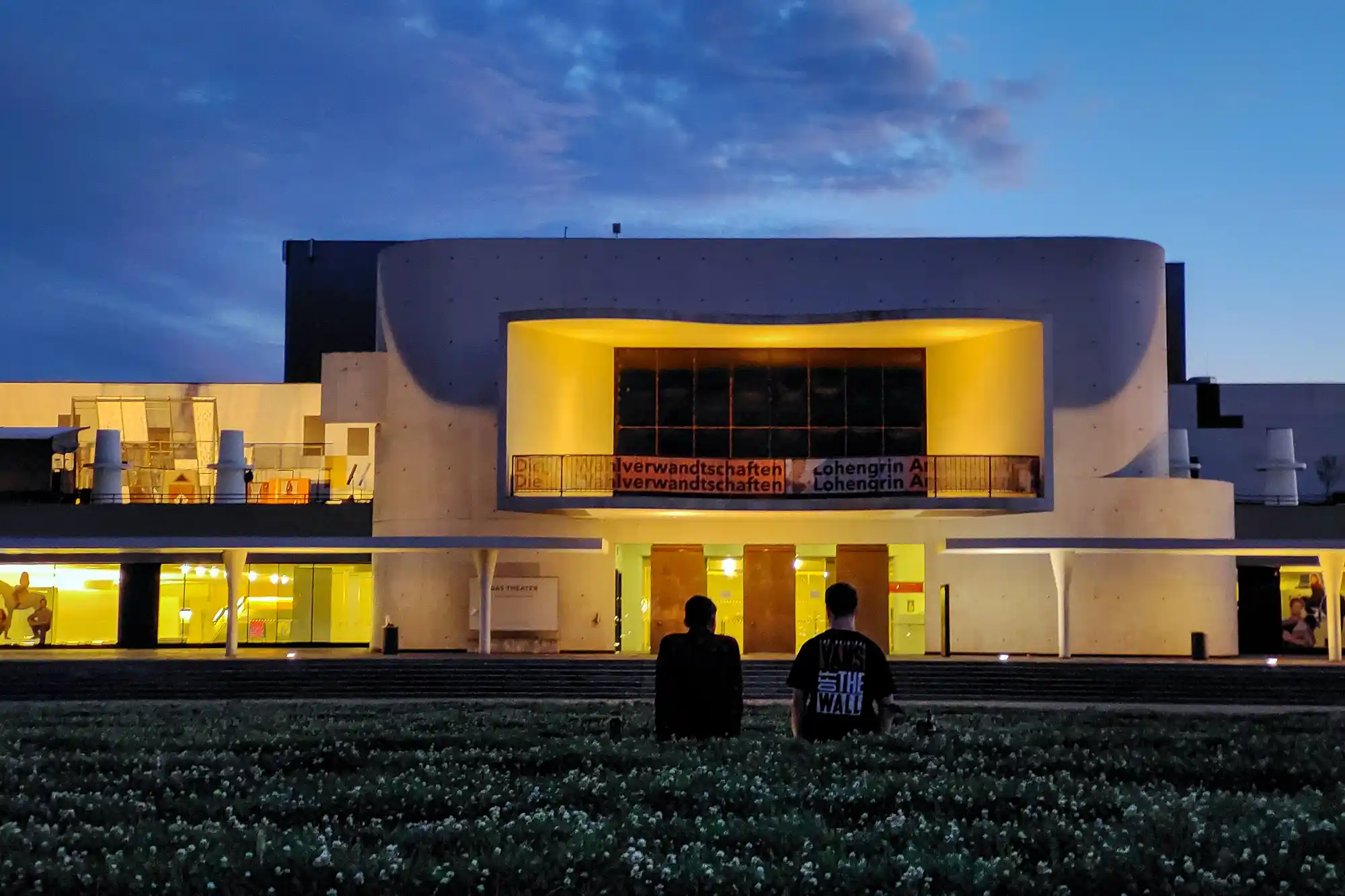 Two people sit on the grass in front of a curved concrete building in Darmstadt, glowing yellow under a blue evening sky.