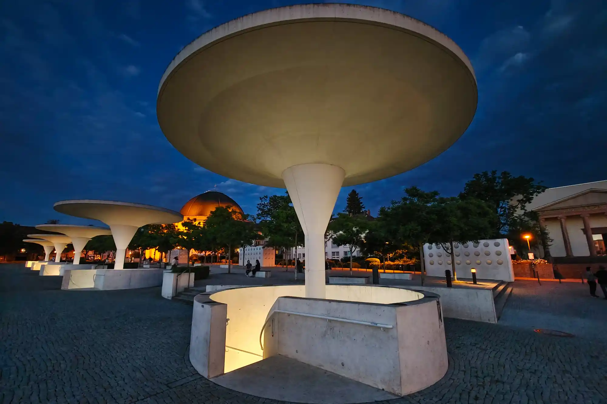 A large pale concrete disc glows under a dark blue sky in Darmstadt, with warm light at its base and a few people nearby.