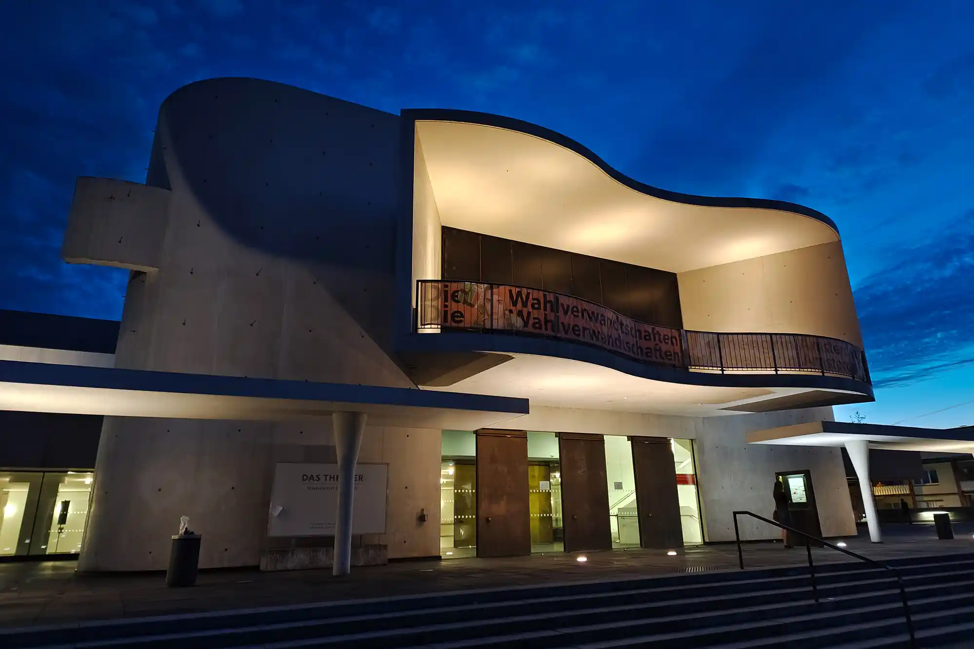 A curved concrete building in Darmstadt glows with warm light under a deep blue sky, with a dark railing curving above the steps.