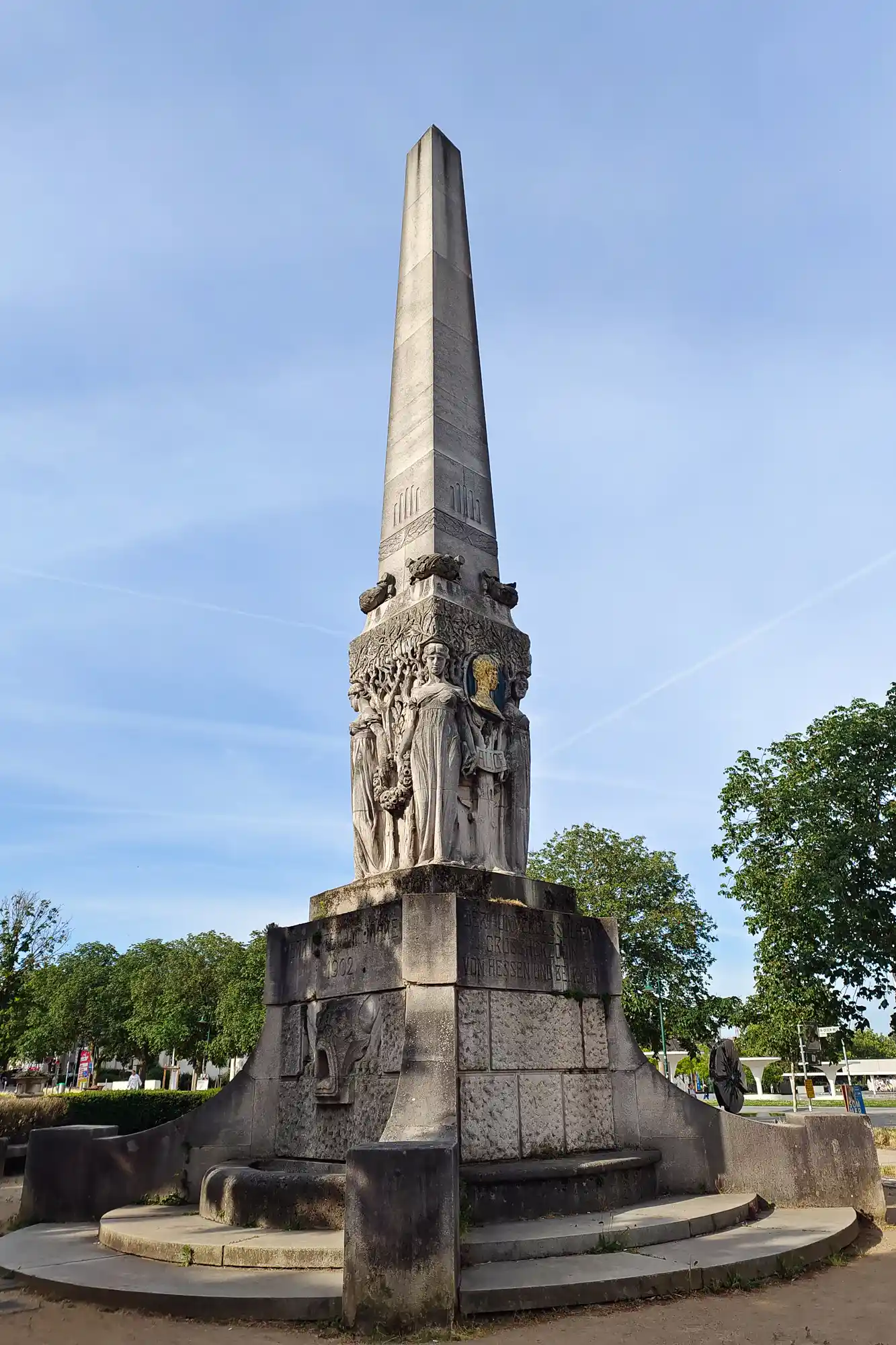 A grey stone obelisk with carved figures in Darmstadt in bright daylight, with a gold emblem and green trees around it.