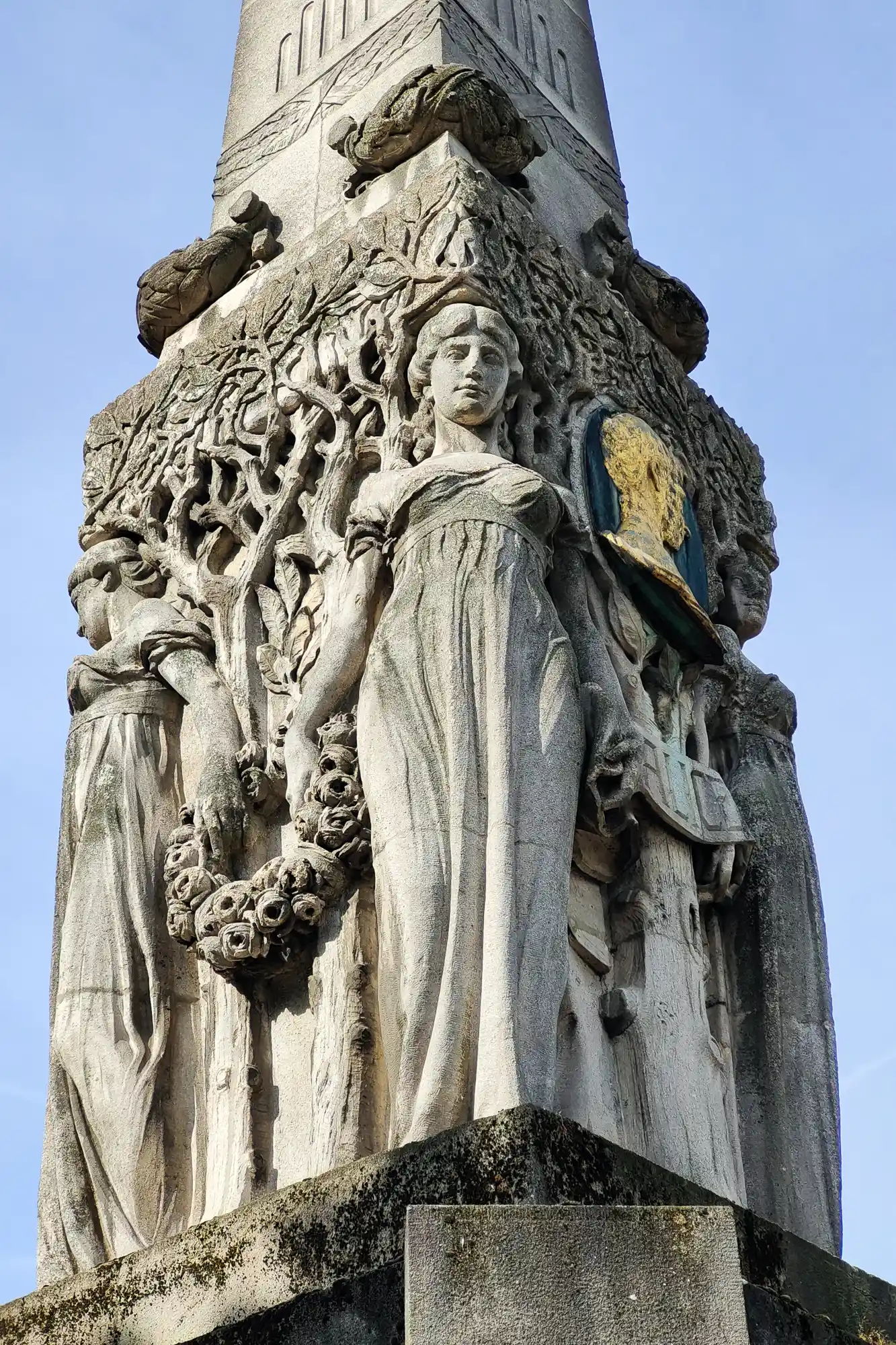 Low-angle view of grey stone figures on a Darmstadt monument, one holding a gold and blue shield, with carved trees behind.