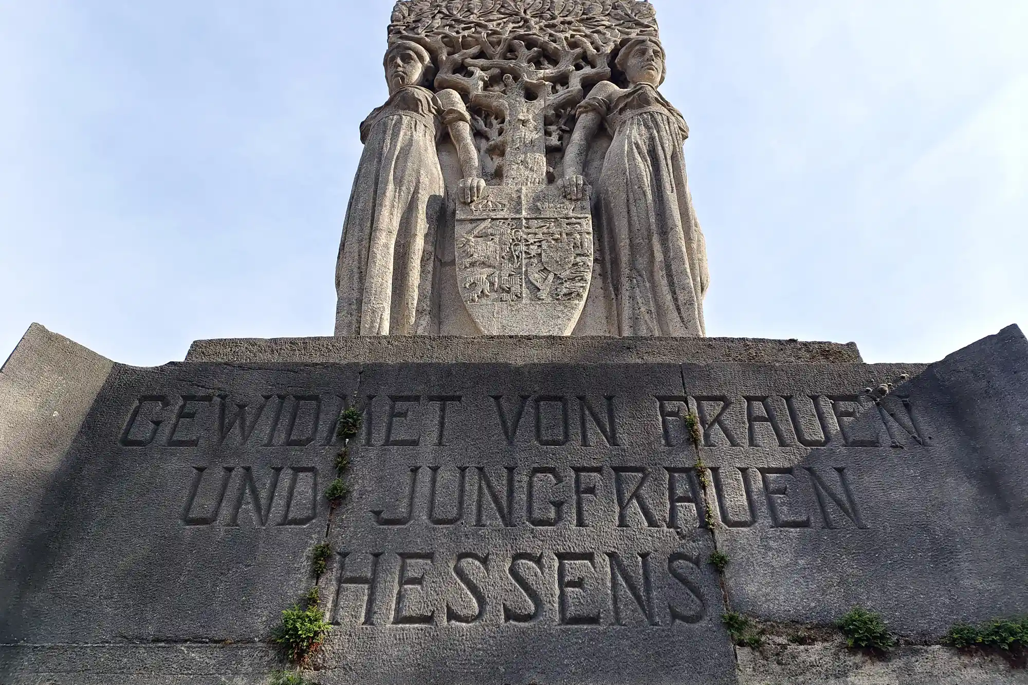 Two grey stone figures holding a shield on a Darmstadt monument, seen from below with green plants growing on the base.
