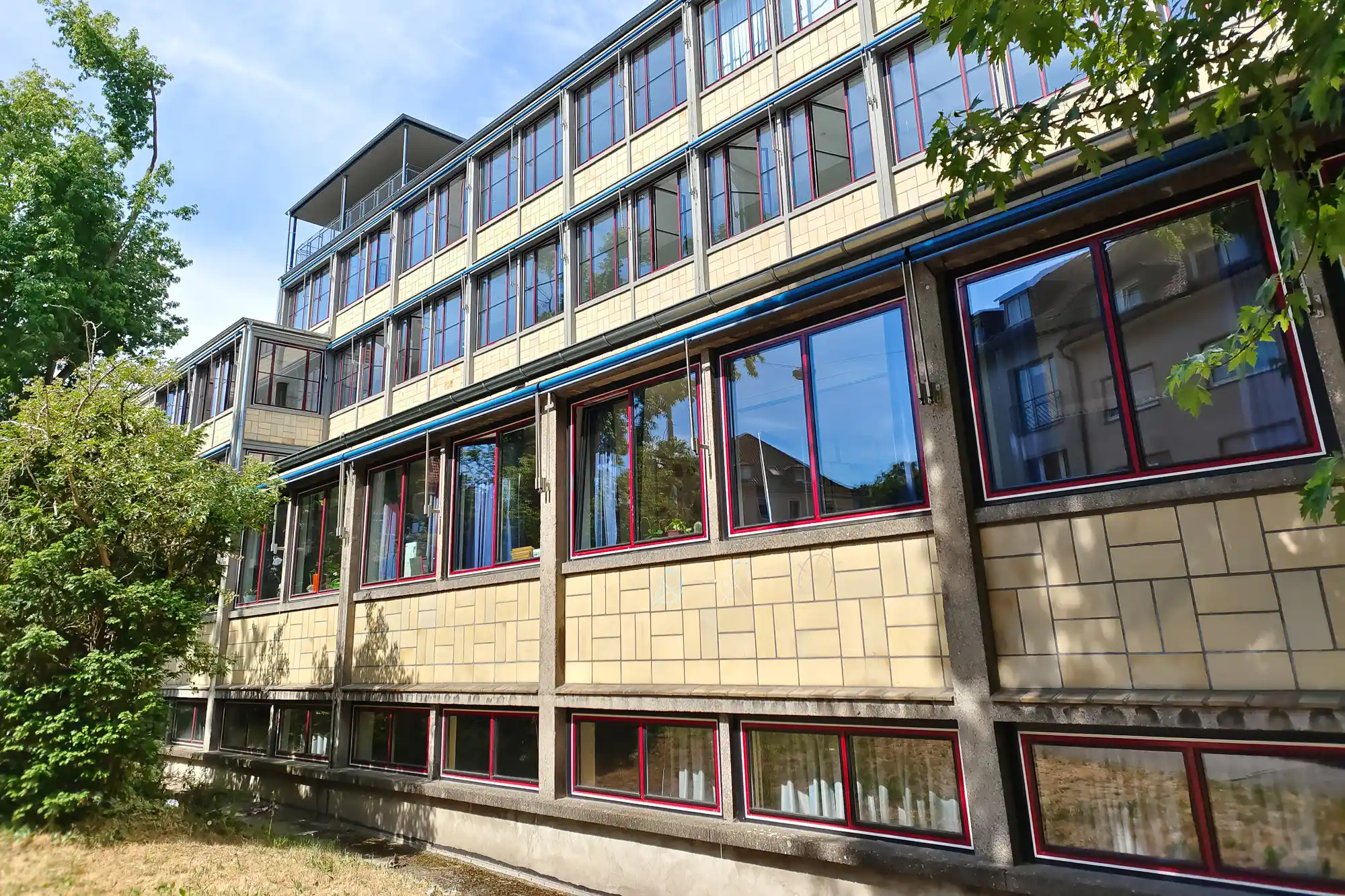 A tan brick building with red-framed windows in Darmstadt in bright sunlight, with green trees casting shadows across it.