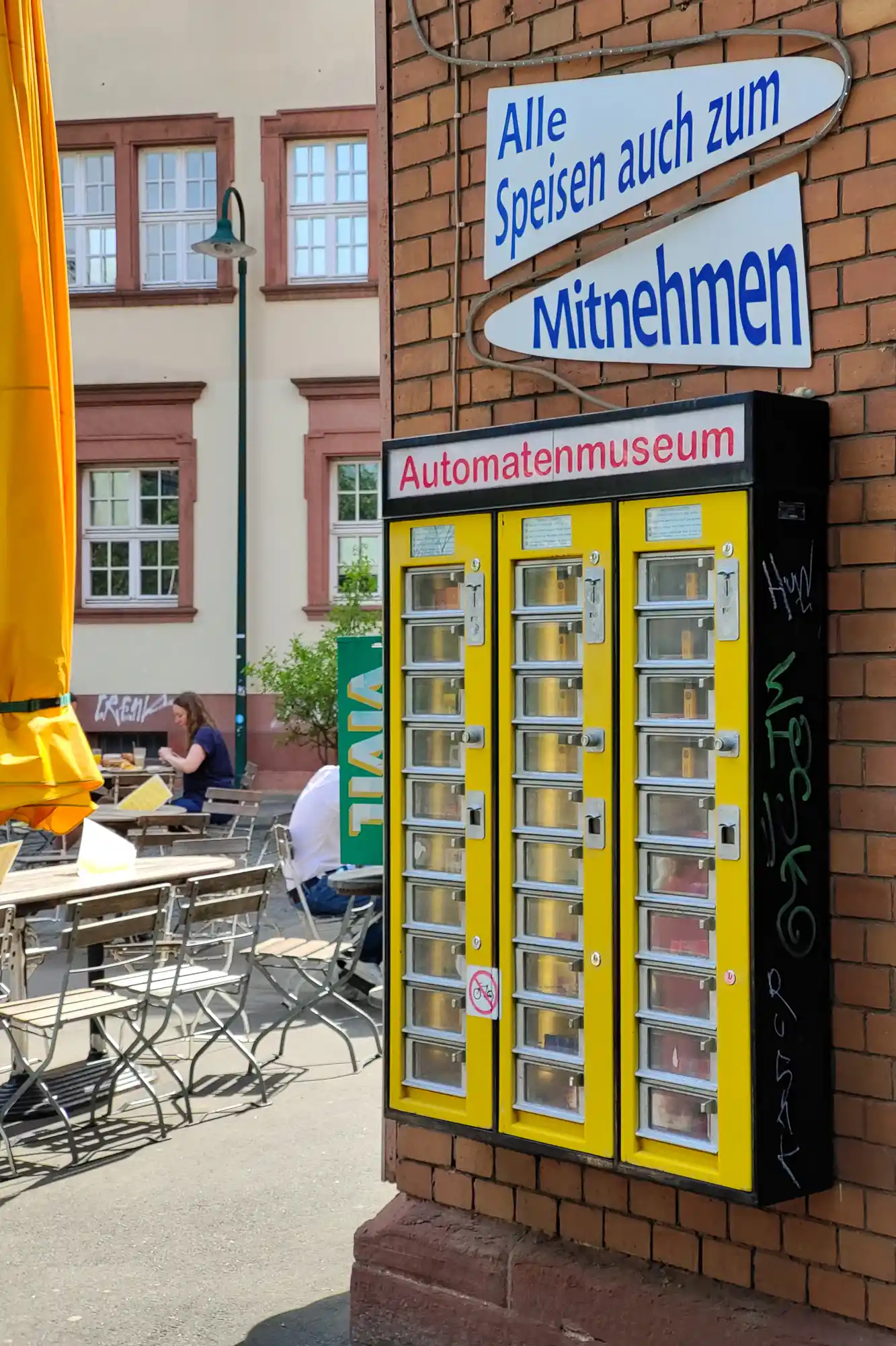 Yellow vending machines with white signs on a brick wall in Darmstadt, beside a yellow umbrella and people seated in the sun.