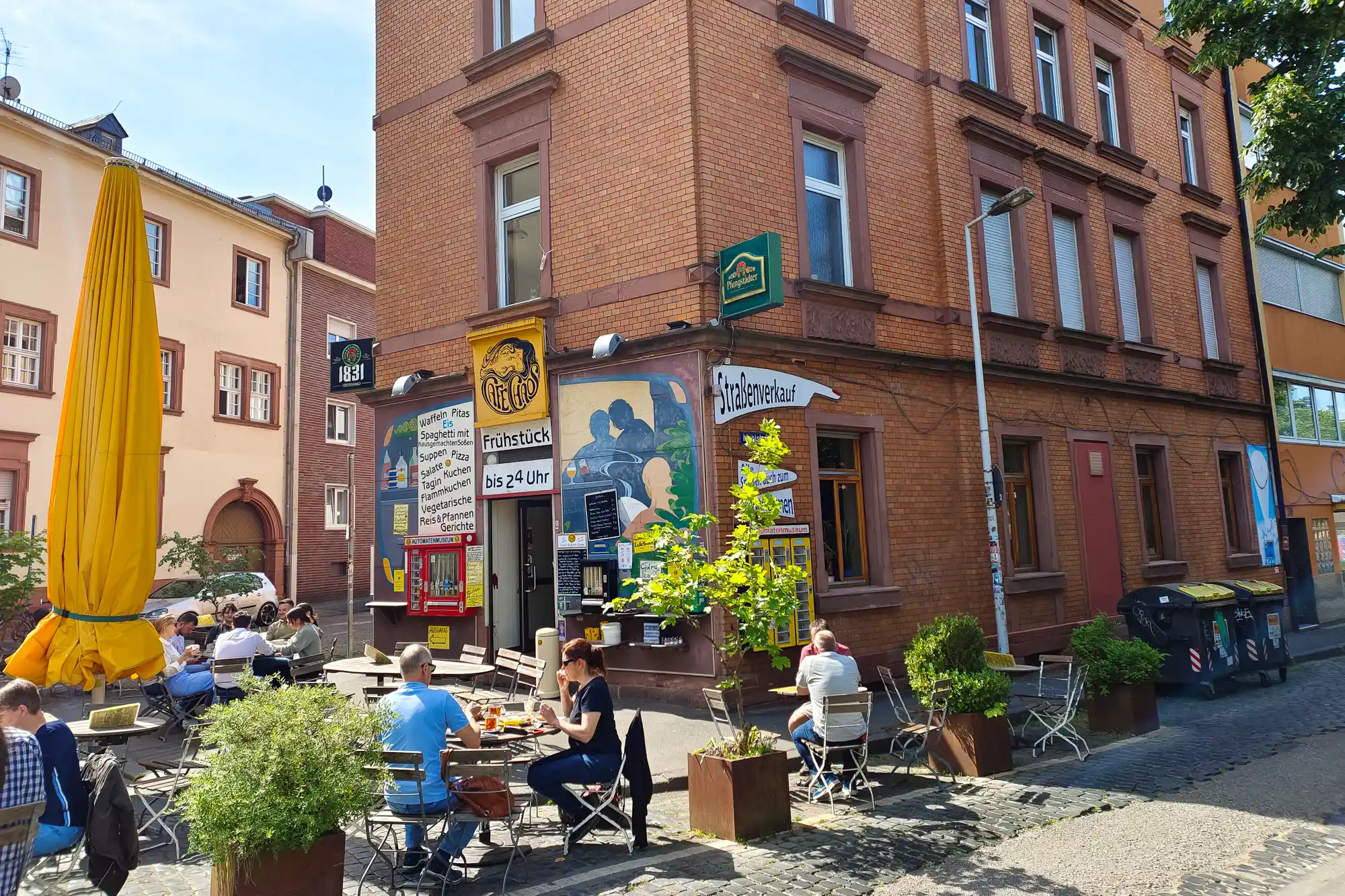 People sitting at tables beside a brick building in Darmstadt, under a yellow umbrella among green plants in the sunshine.