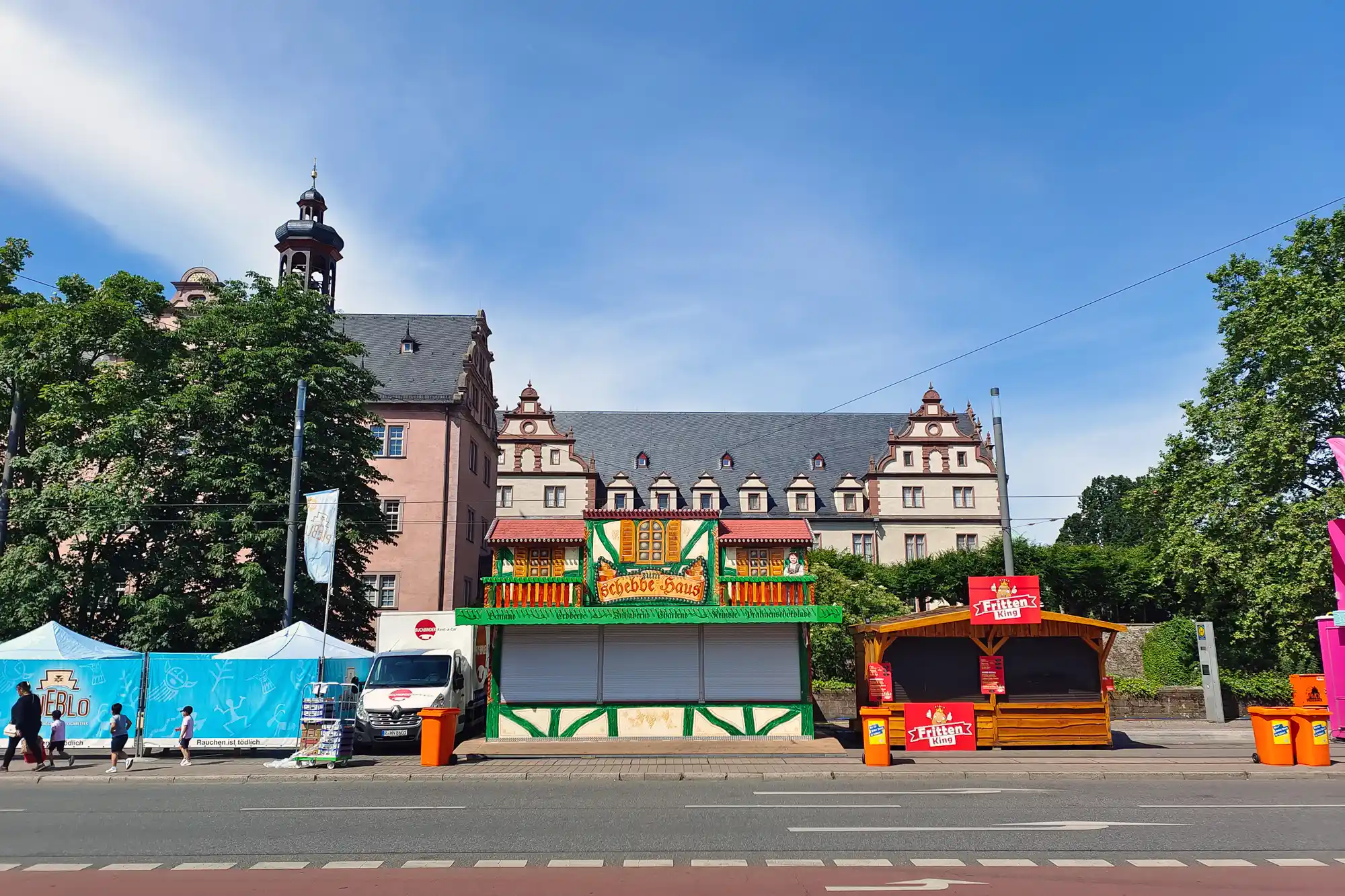 A green and yellow stall with red trim in bright light in Darmstadt, with people walking past blue fabric and orange bins.