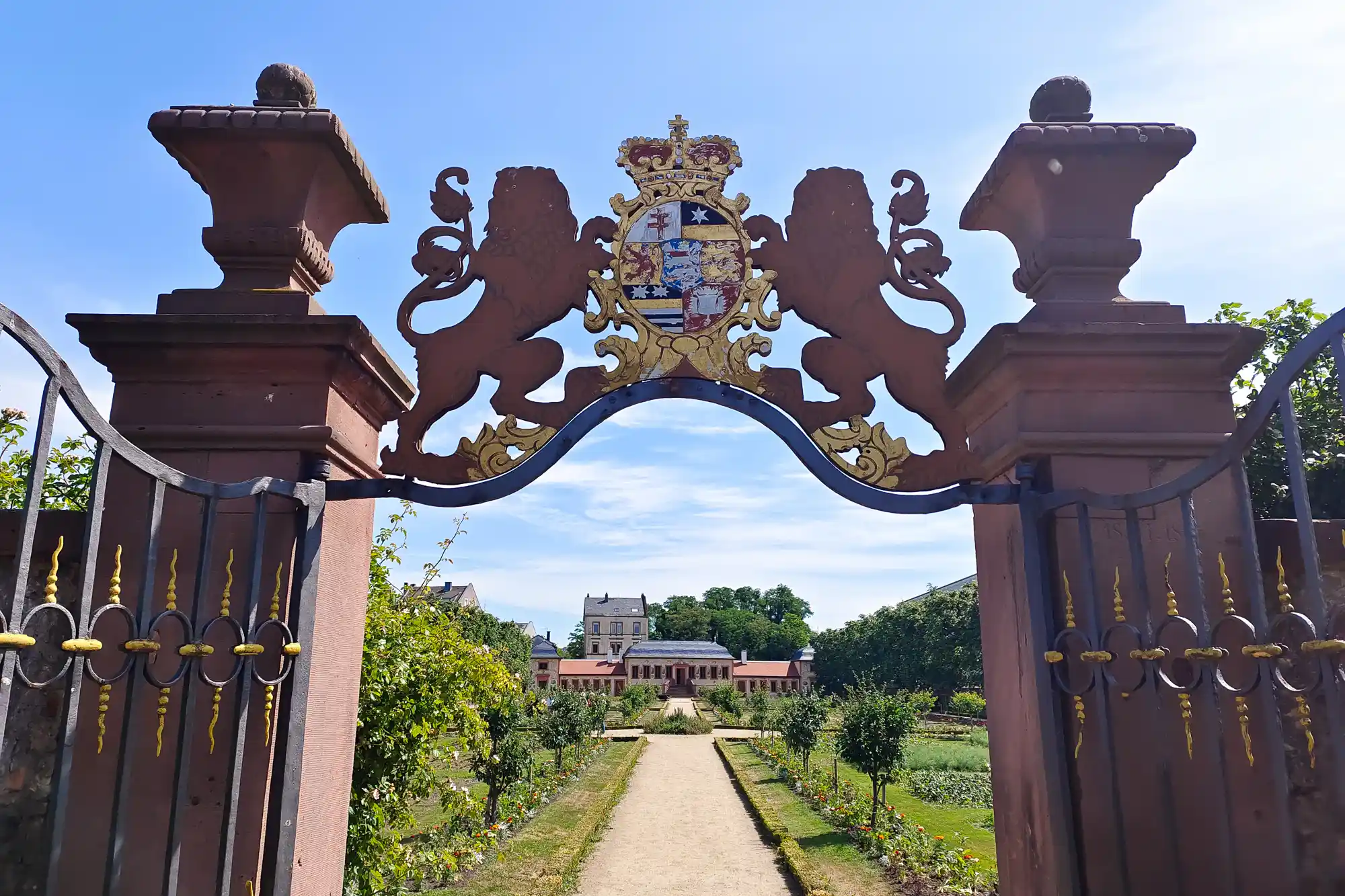 A reddish-brown gate with gold accents and a crest in Darmstadt's Herrengarten, opening onto a sunlit path through green plants.