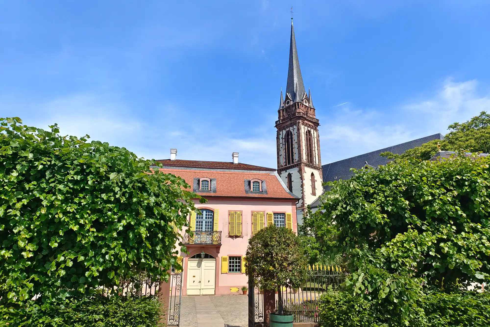 A pink building with yellow shutters and a red roof behind green trees in Darmstadt, with a tall dark spire rising in bright light.