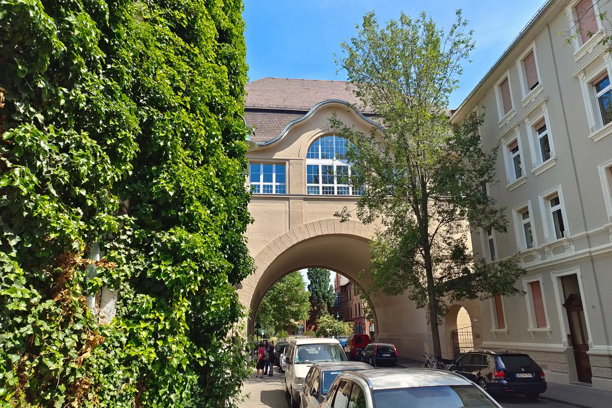 A tan archway with a curved roof and glass windows in Darmstadt, framed by green foliage and a row of parked cars.