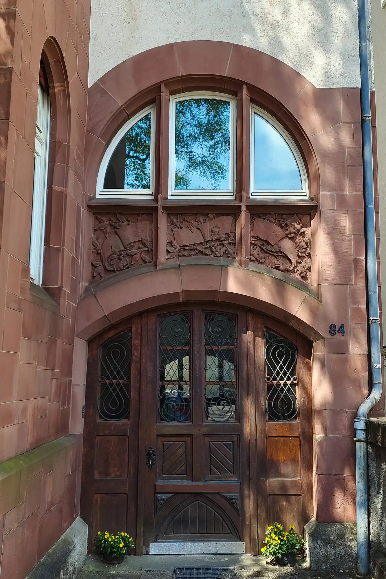 A carved reddish stone arch around a dark wooden door with metal scrollwork in Darmstadt, with yellow flowers below in the sun.