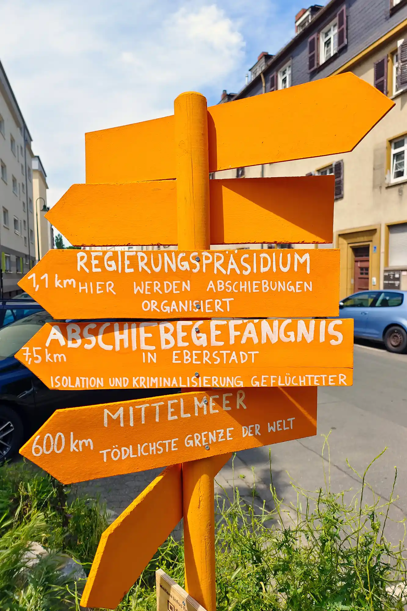 Bright yellow wooden signs with white lettering in Darmstadt, lit by the summer sun above green plants.