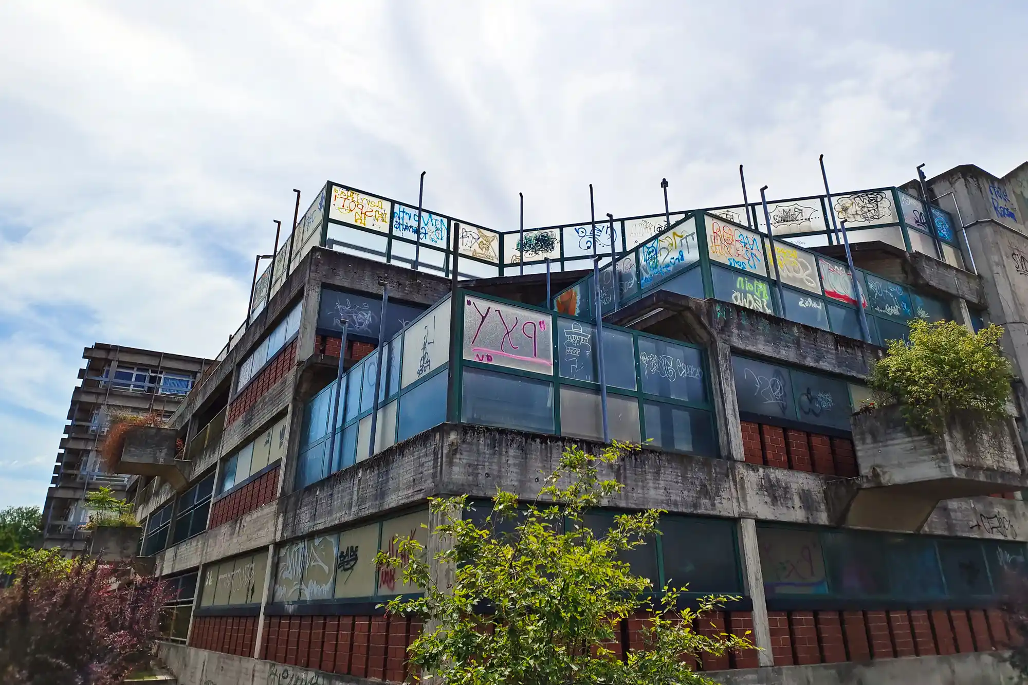 A concrete building in Darmstadt with graffiti on its glass panels and red brick sections, with green plants growing from it.