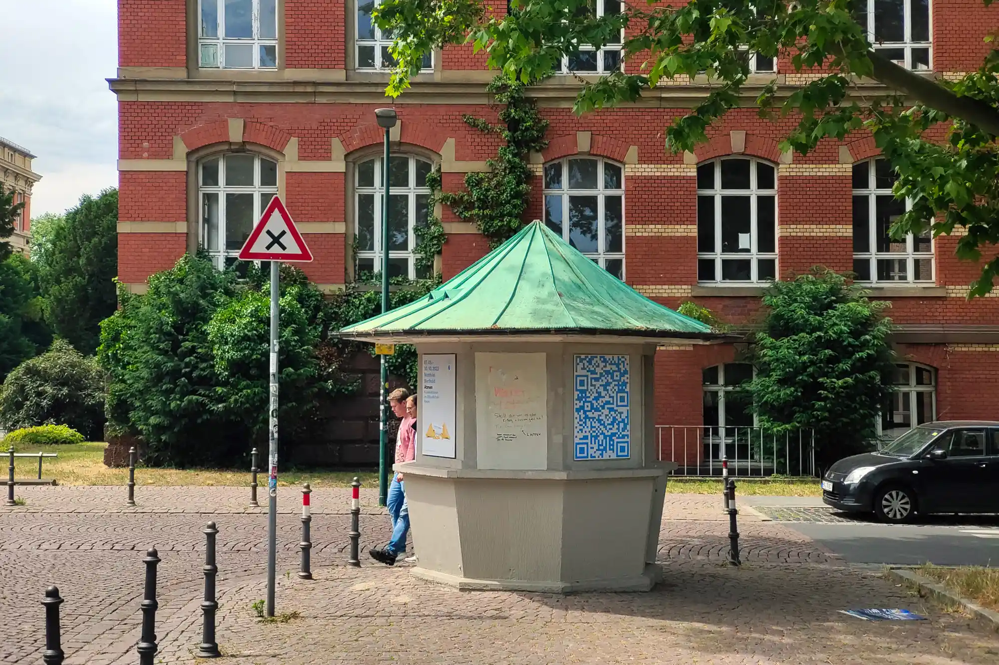A grey kiosk with a green roof on cobblestones in Darmstadt, with two people walking past red brick walls.